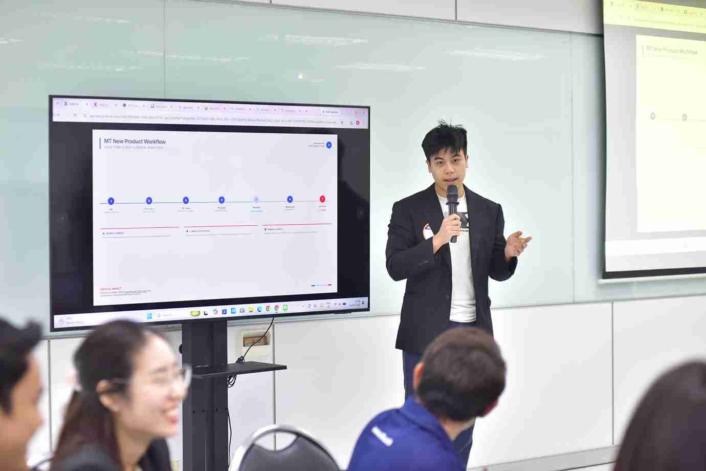 Man presenting with microphone, timeline displayed on screen, audience watching.