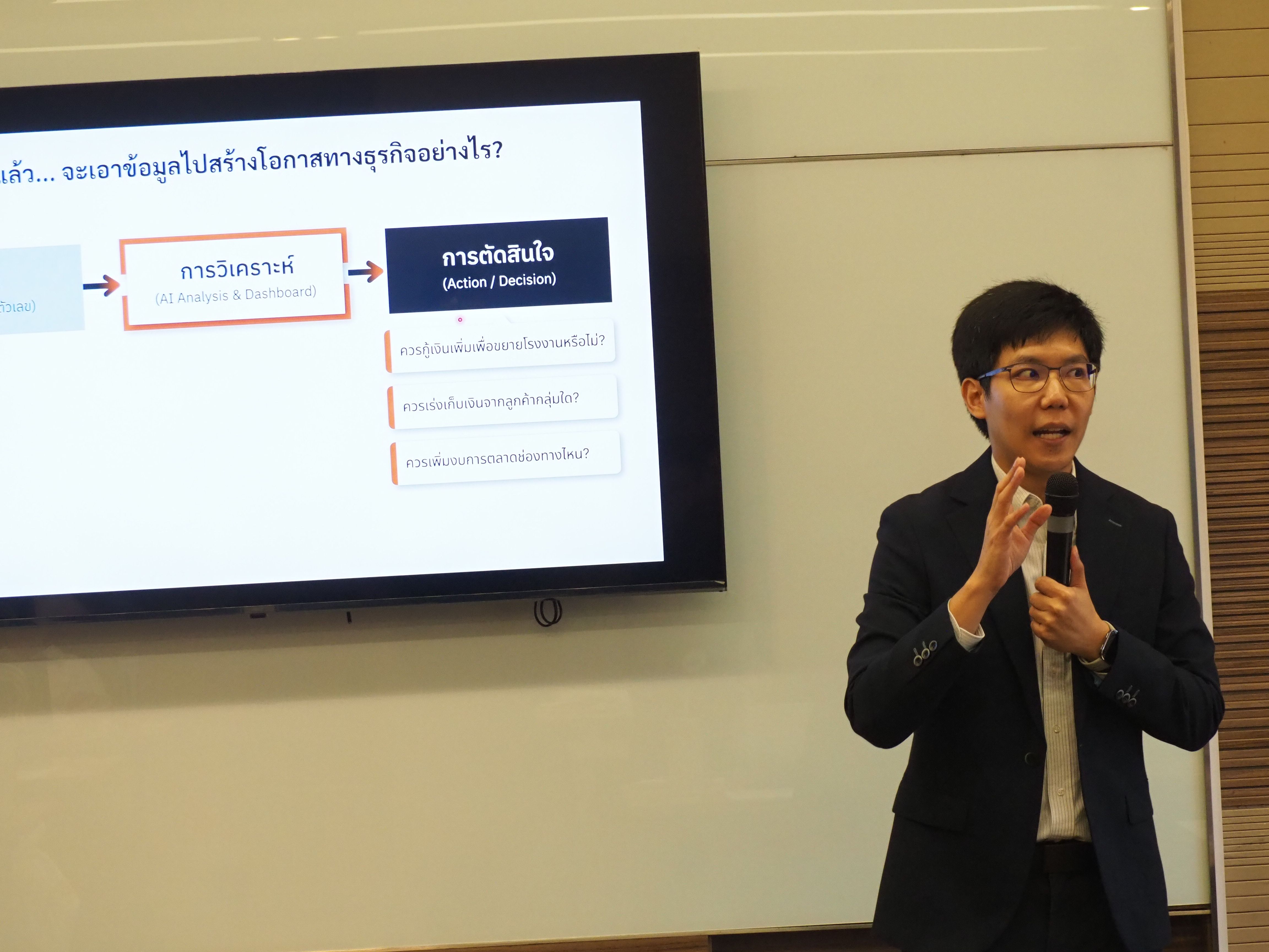 Man in suit presenting next to a screen with a Thai flowchart diagram.