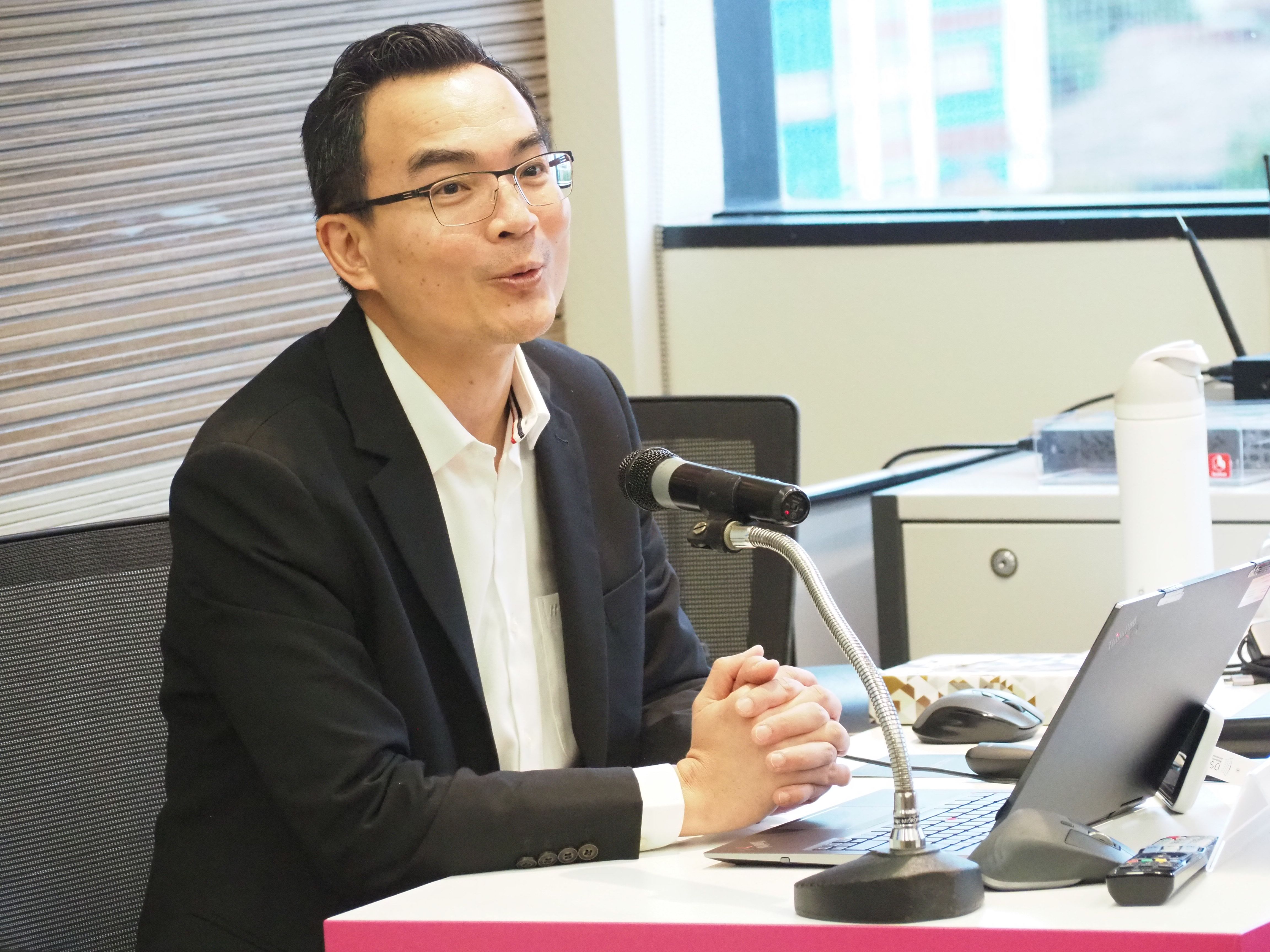 A man in glasses and a dark jacket speaks into a microphone at a table.