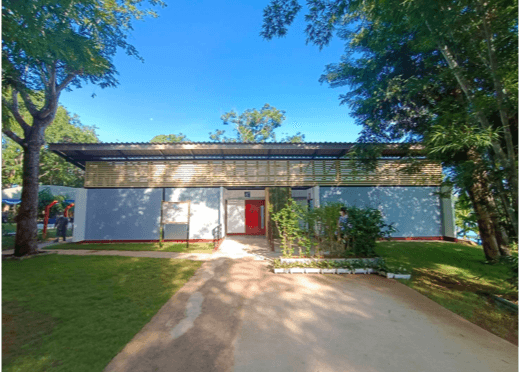 Modern building with gray walls, a red door, and flat roof, surrounded by green trees.