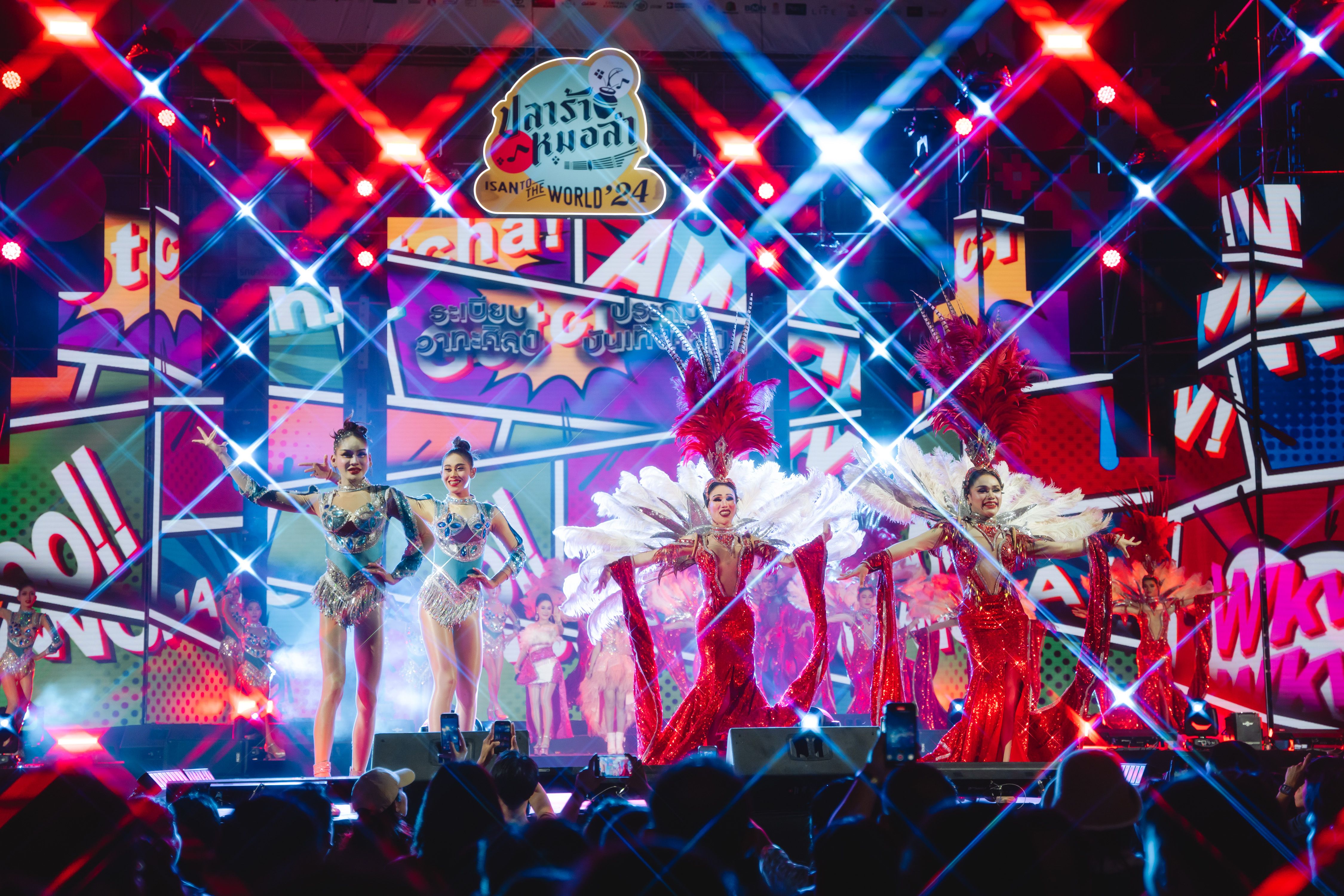 Performers in vibrant costumes dance on a brightly lit stage with colorful pop art graphics.