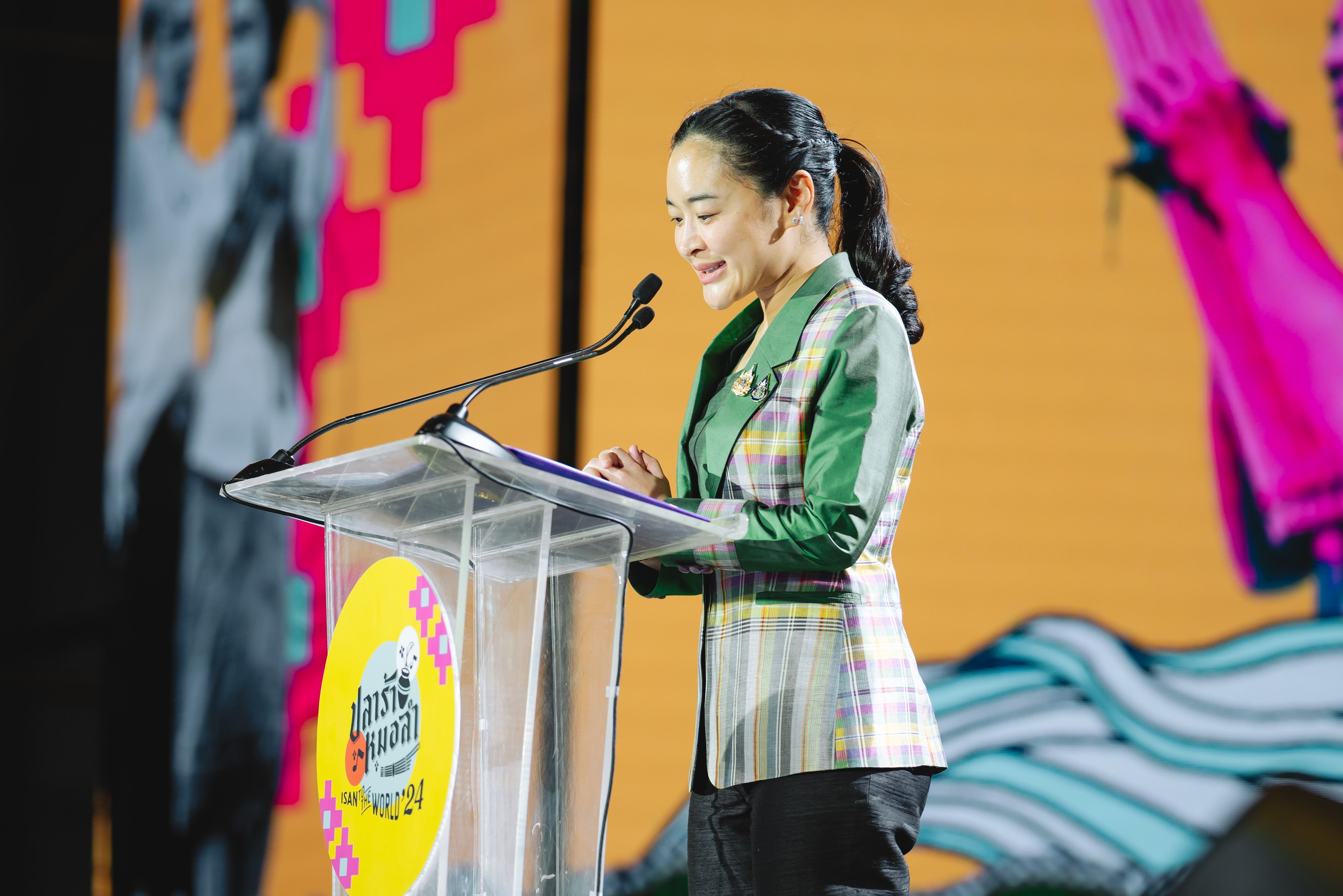 Woman in plaid jacket speaks at clear podium against a colorful background.