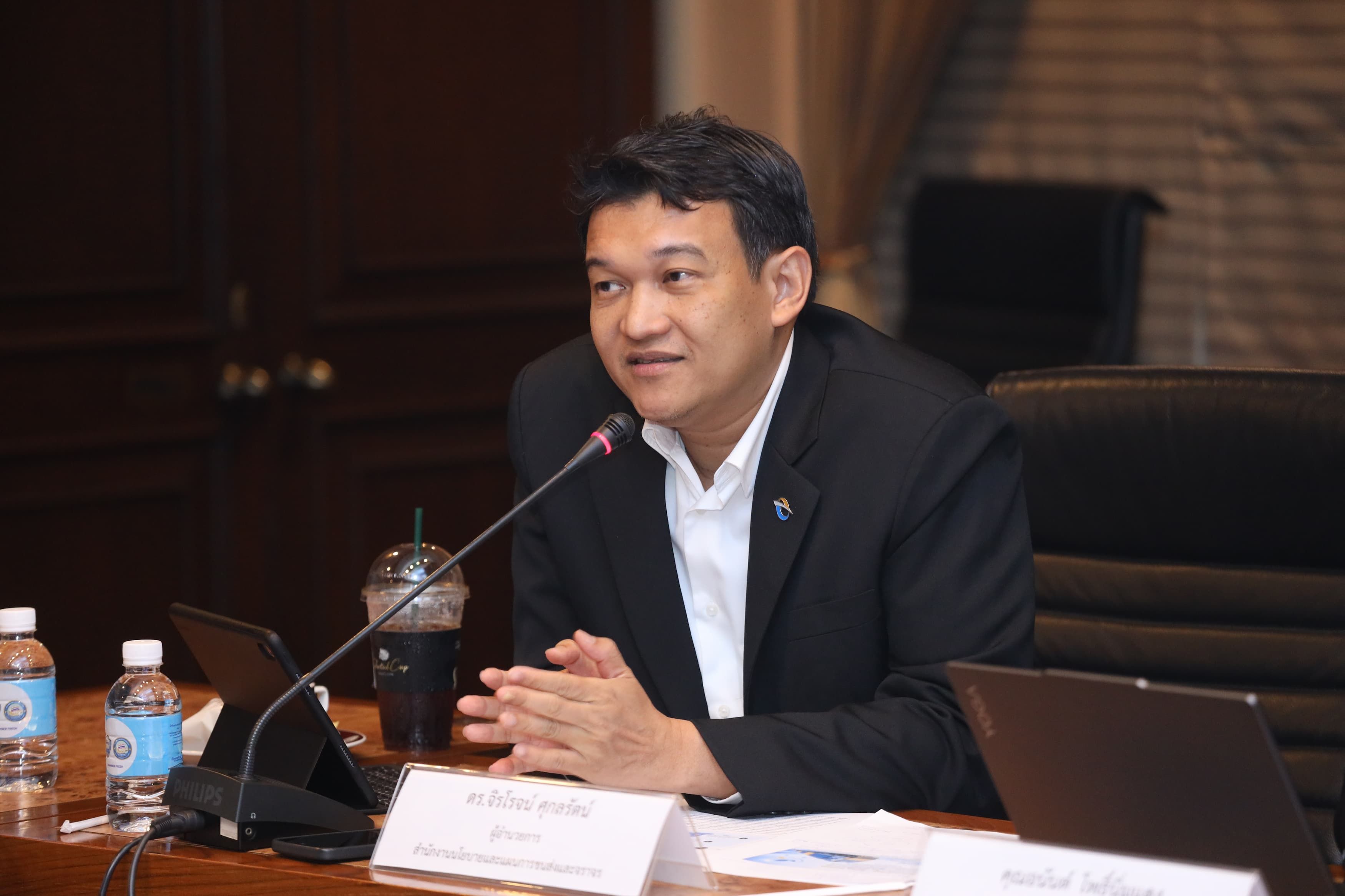 An Asian man in a dark suit speaks into a microphone at a conference table.