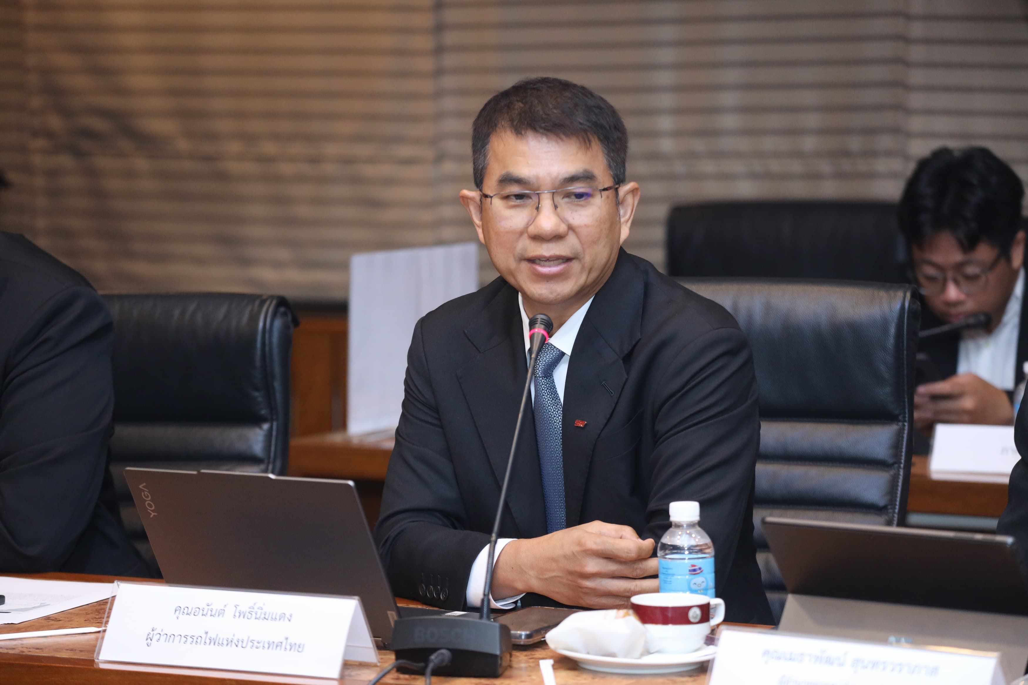 Man in suit and glasses speaking at a conference table with a laptop.