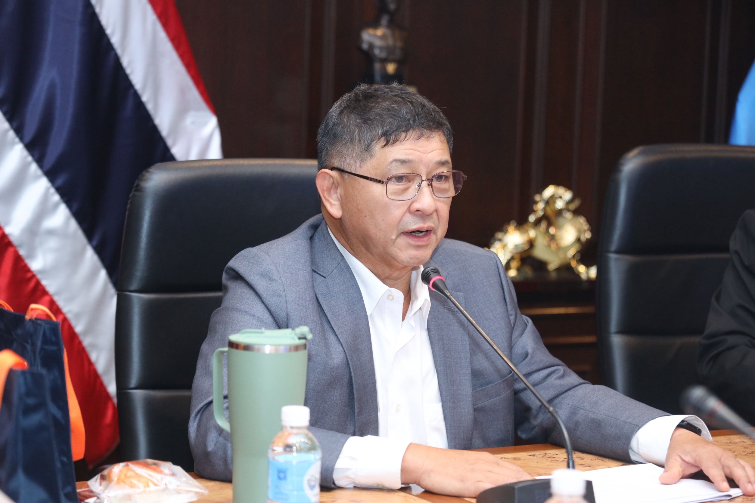 Man in gray suit and glasses speaks into a microphone at a formal table.