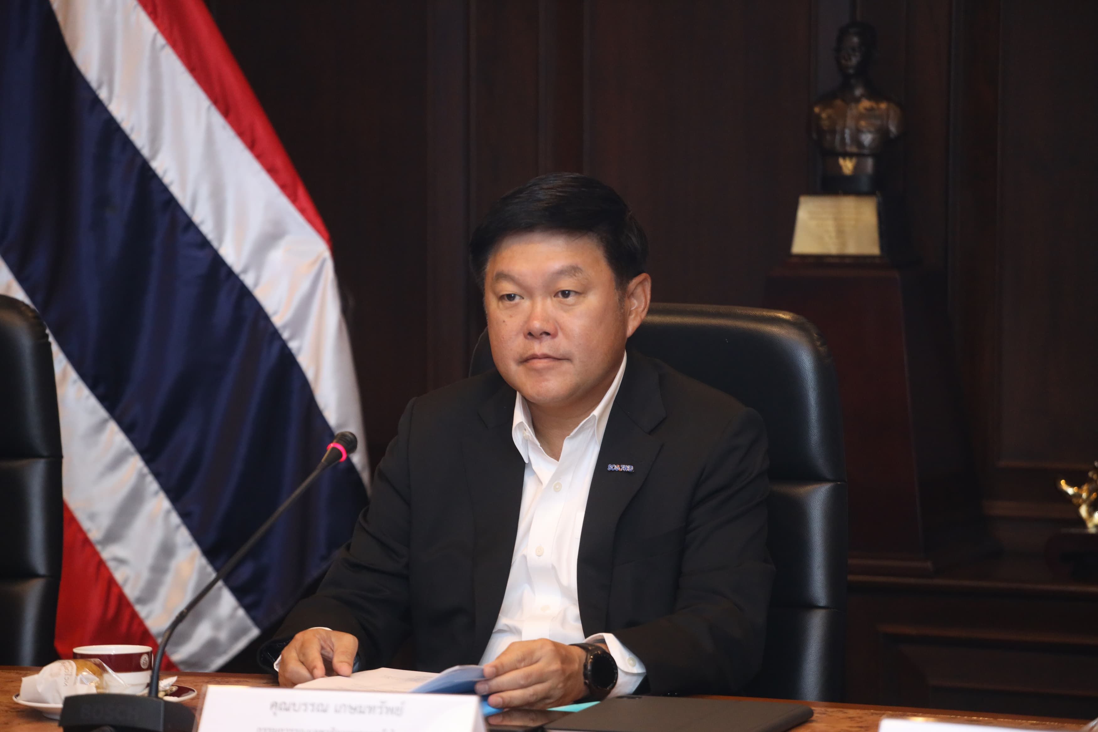 Asian man in a dark suit sits at a desk with Thailand's flag visible.