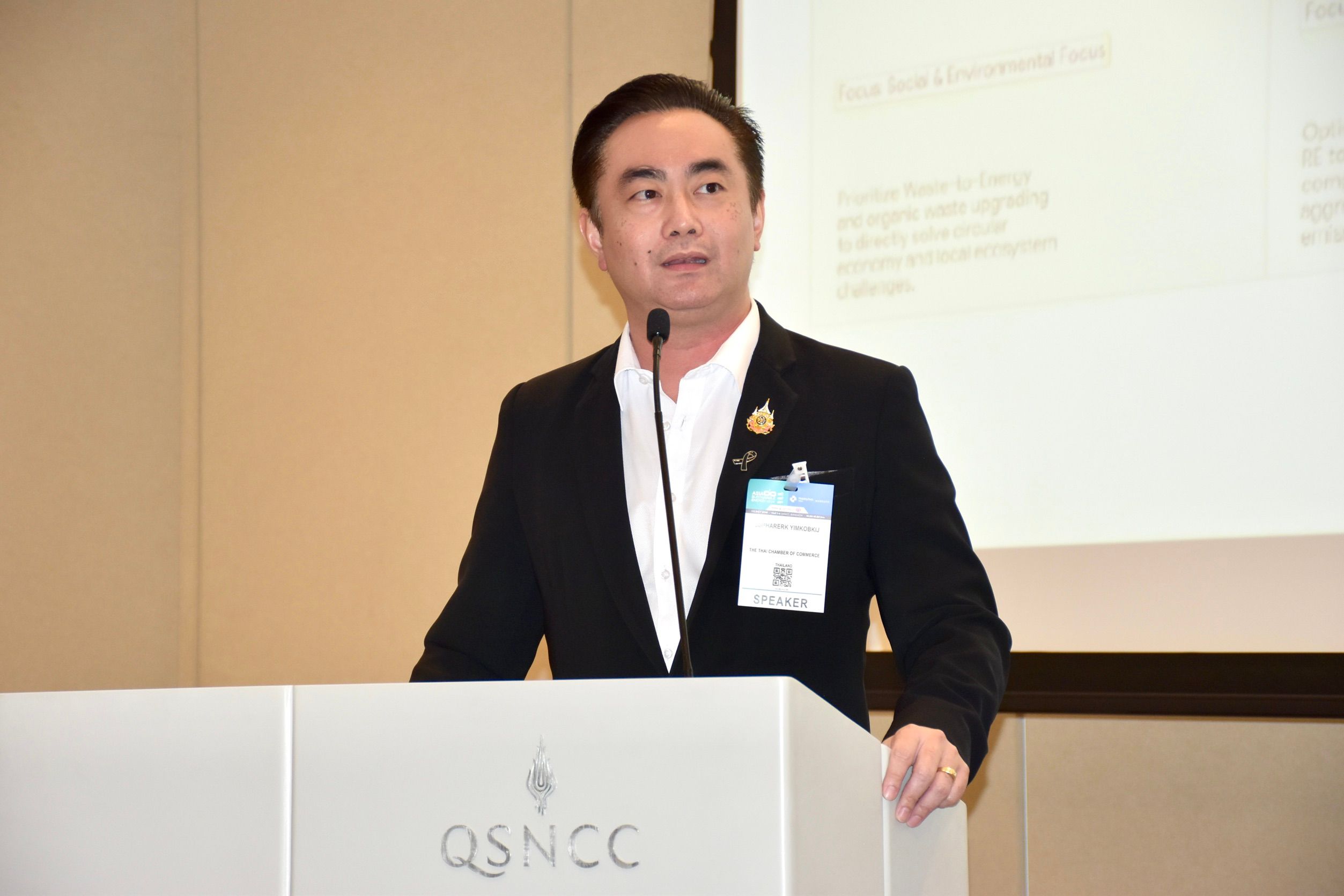A man in a black suit speaks from a podium with the QSNCC logo.