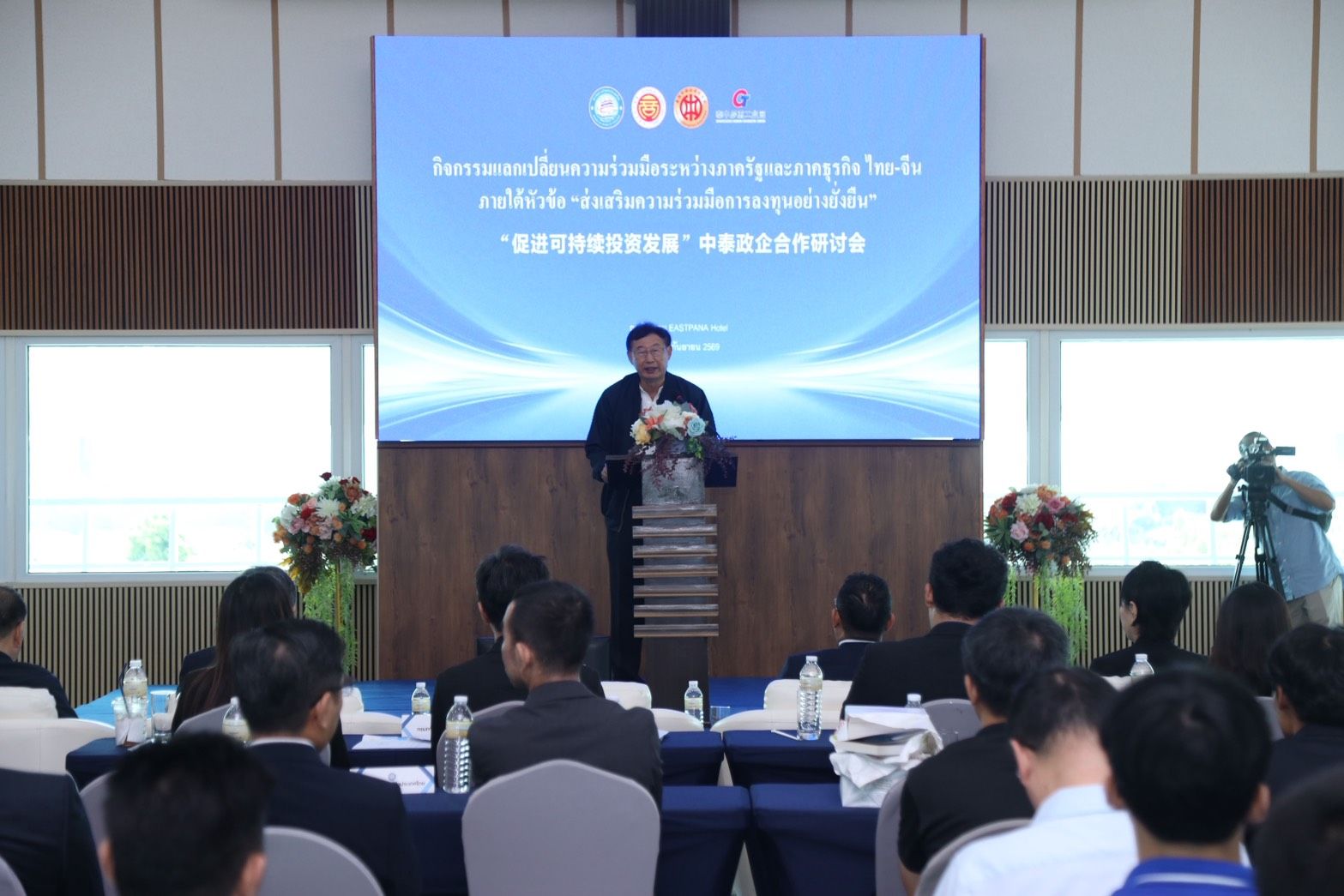 A man delivers a speech from a podium in a conference room to an attentive audience.