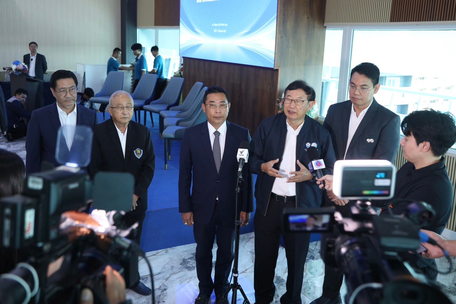 Men in suits speaking at a press conference with microphones and cameras.