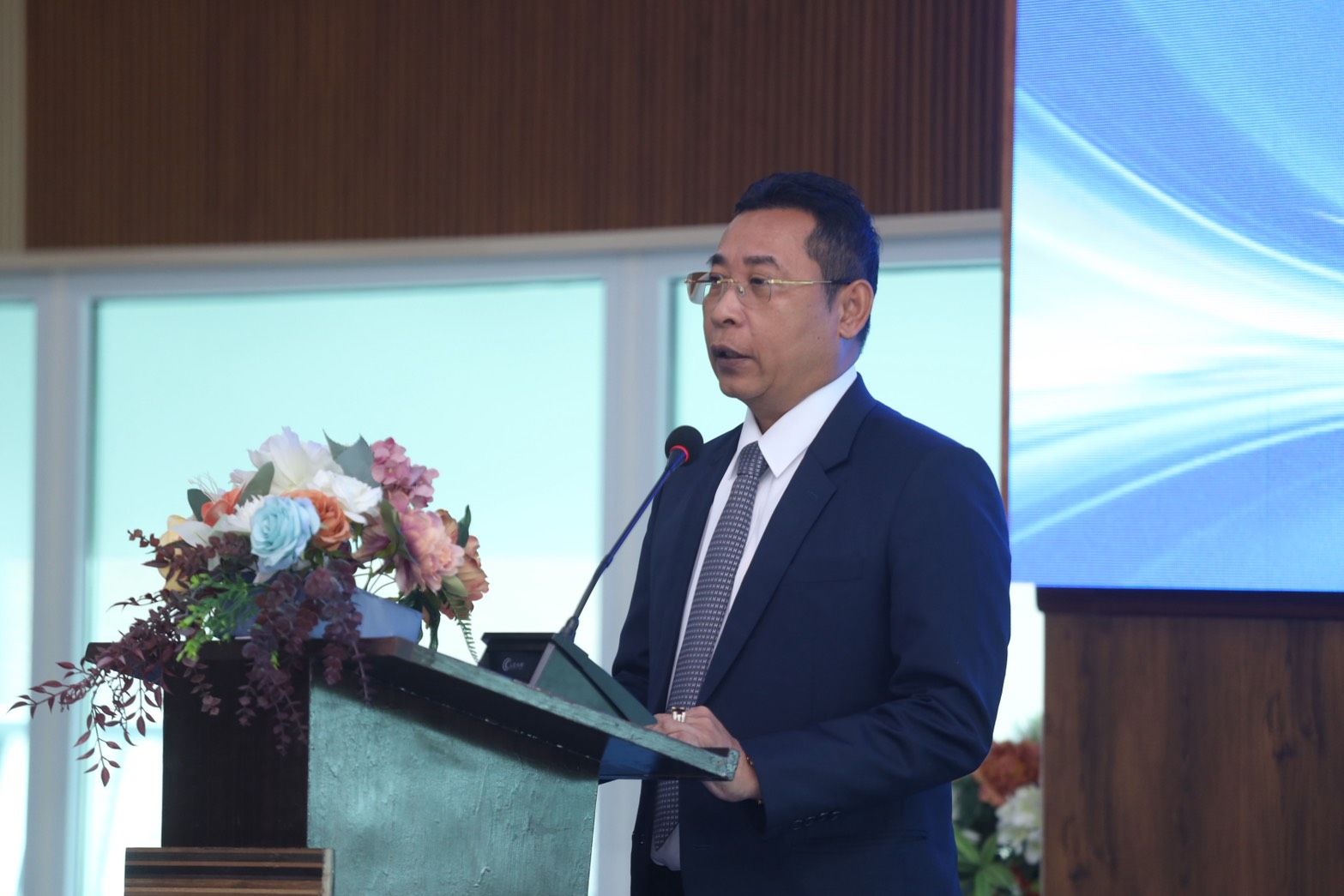 Man in dark suit and glasses speaks at a podium with flowers in a modern room.