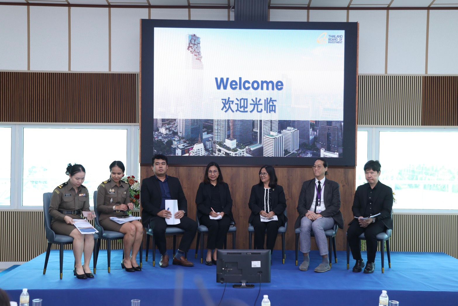 Seven people sit on a panel before a large screen displaying "Welcome" at a conference.