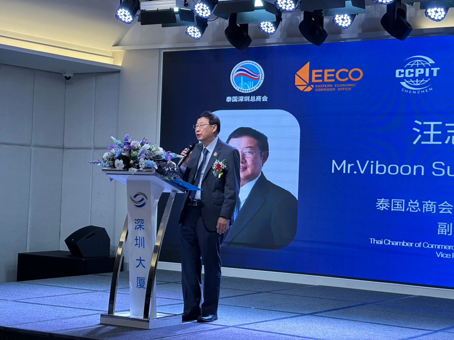 Man in suit speaks at podium with presentation screen showing logos and text.