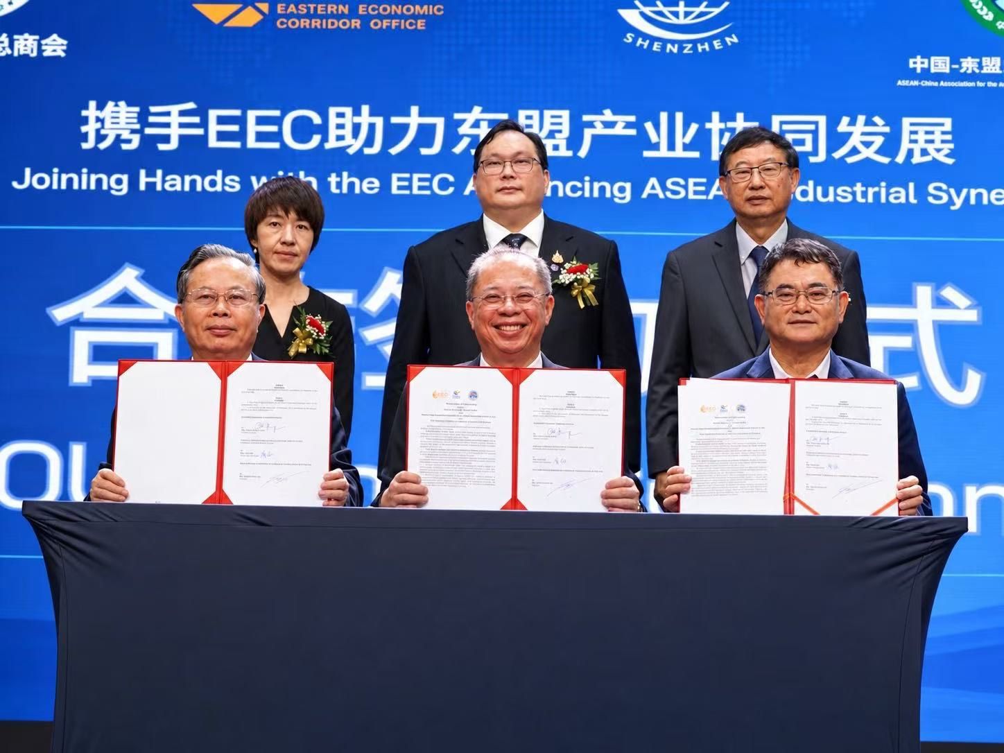 Six officials at a signing ceremony, three seated holding signed agreements.