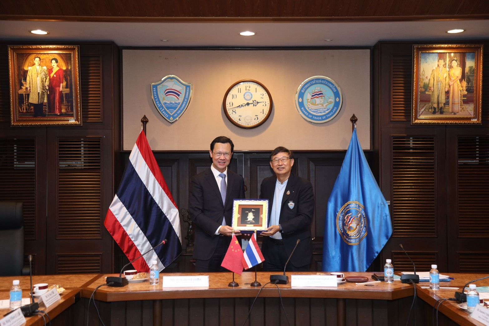 Two men in suits exchange a framed gift, standing before Thai and blue flags.