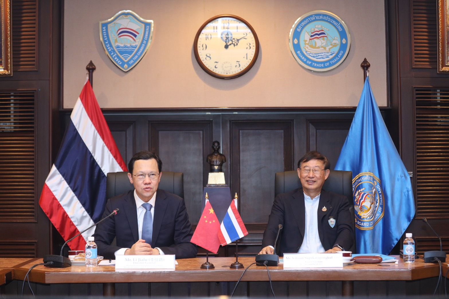 Two men in suits sit at a meeting table with Thai and Chinese flags displayed.