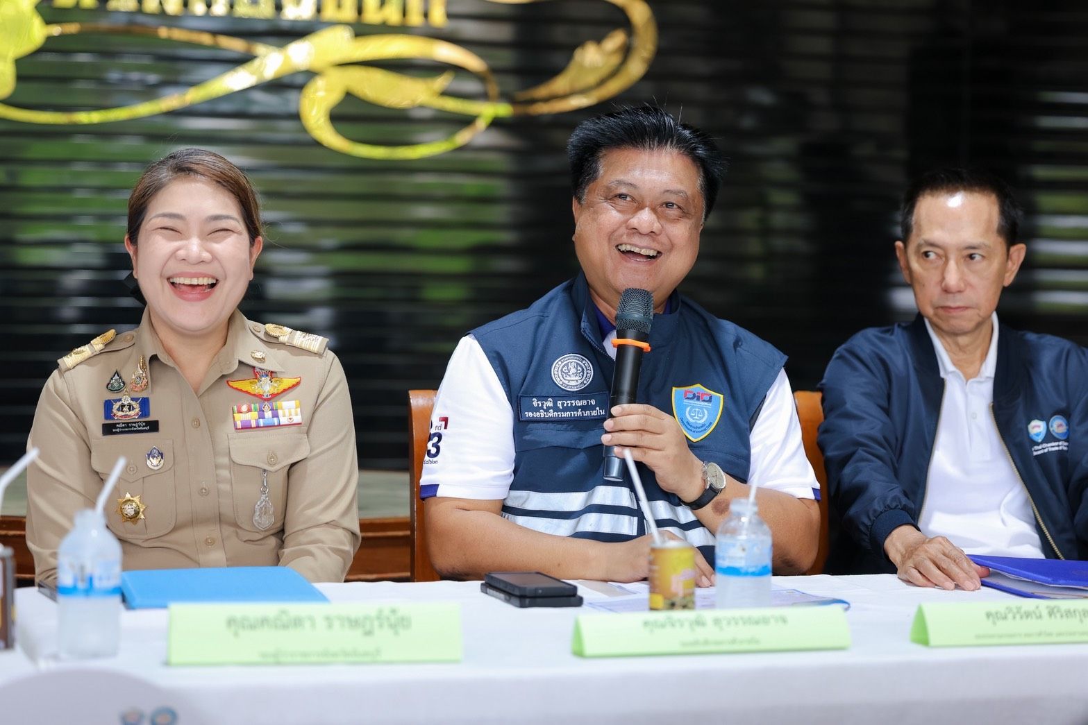 Three officials are seated at a table, two laughing while one man speaks into a microphone.