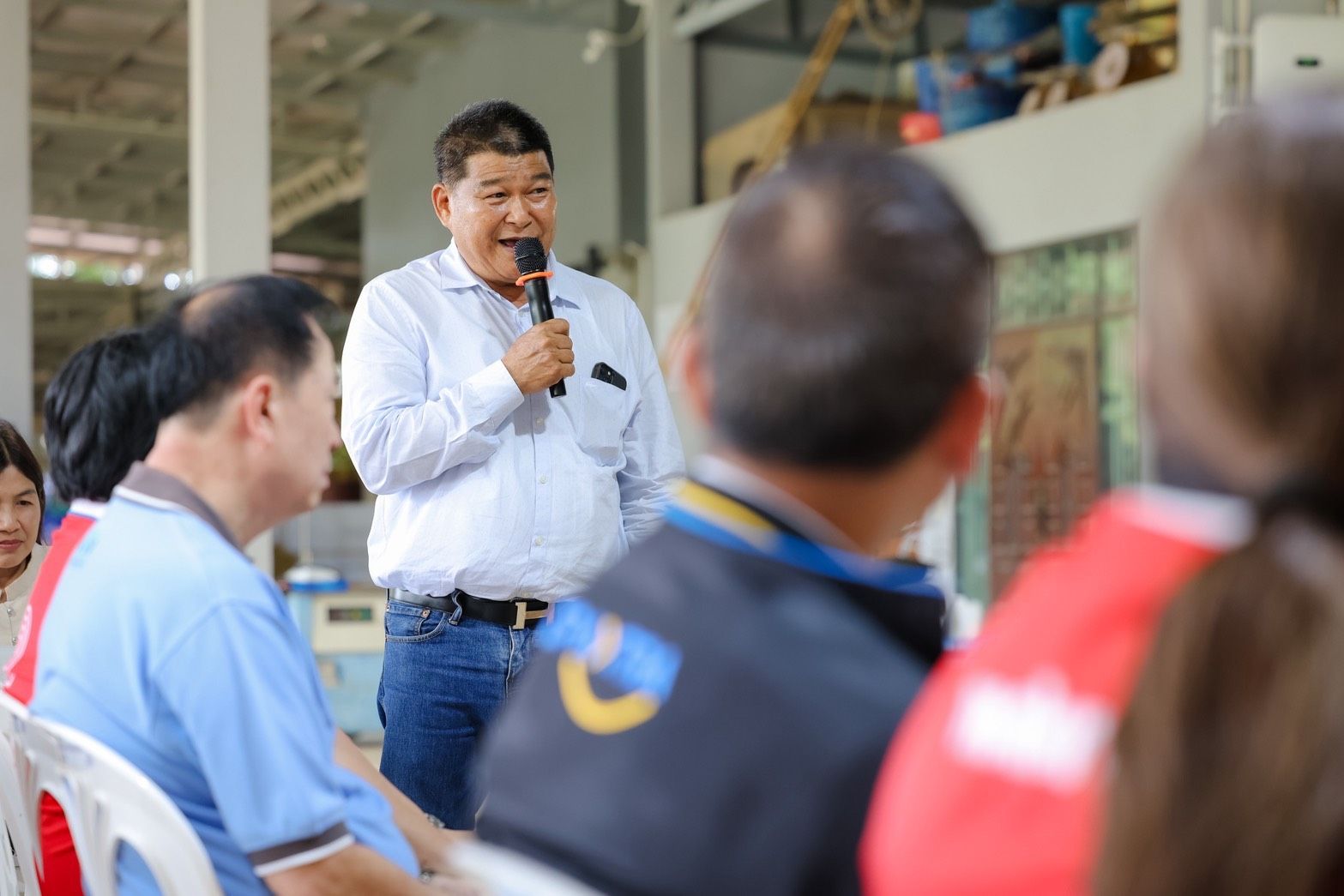 Man in white shirt speaks into microphone addressing a group of people indoors.
