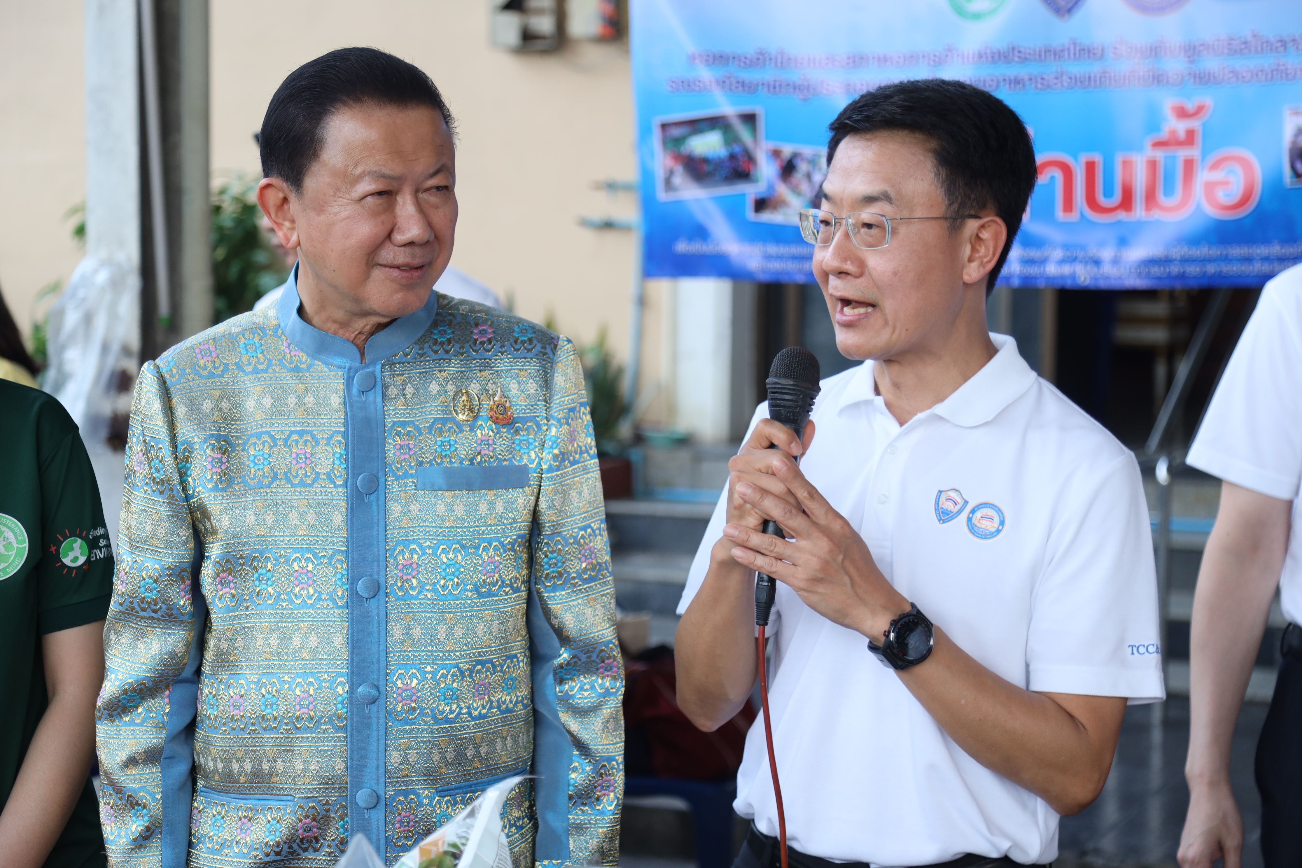 Older man in traditional Thai jacket listens to younger man speaking into a microphone.
