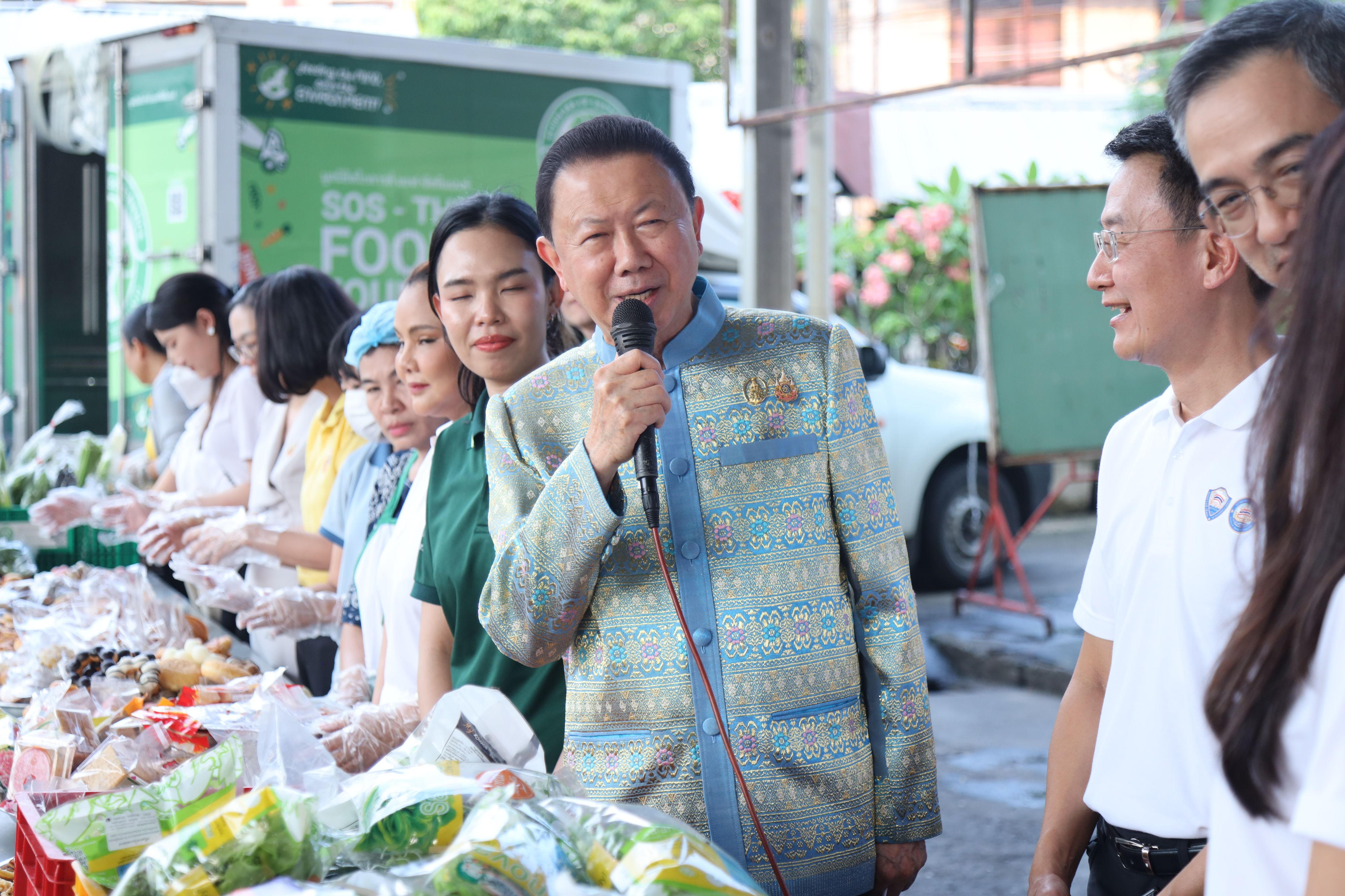 Man in traditional attire speaking into microphone at an outdoor food distribution event.