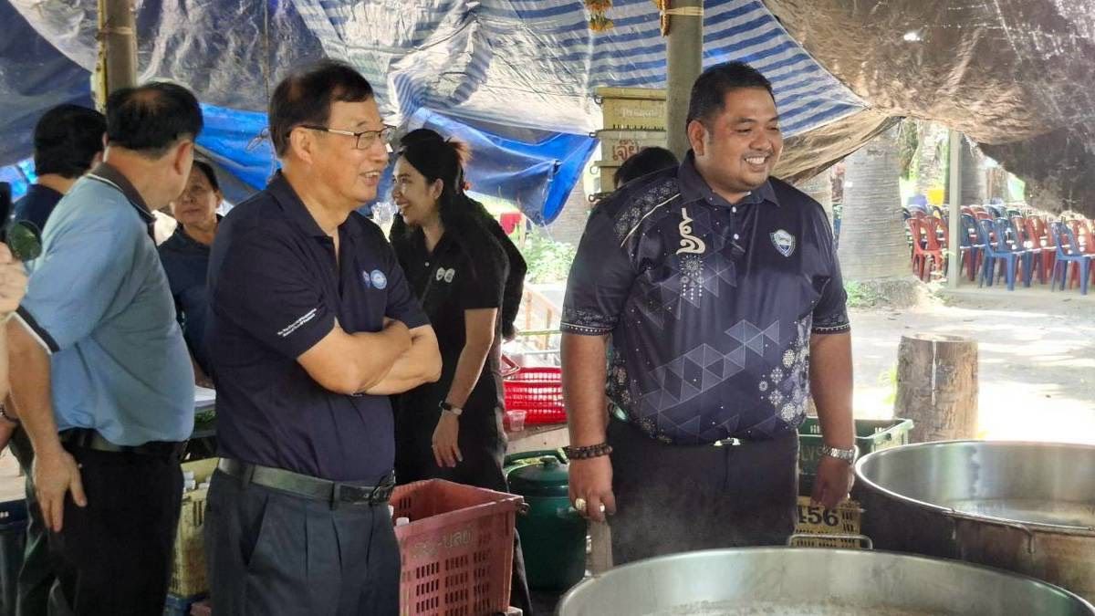 Two men in dark uniform shirts stand next to large cooking pots under a tarp.