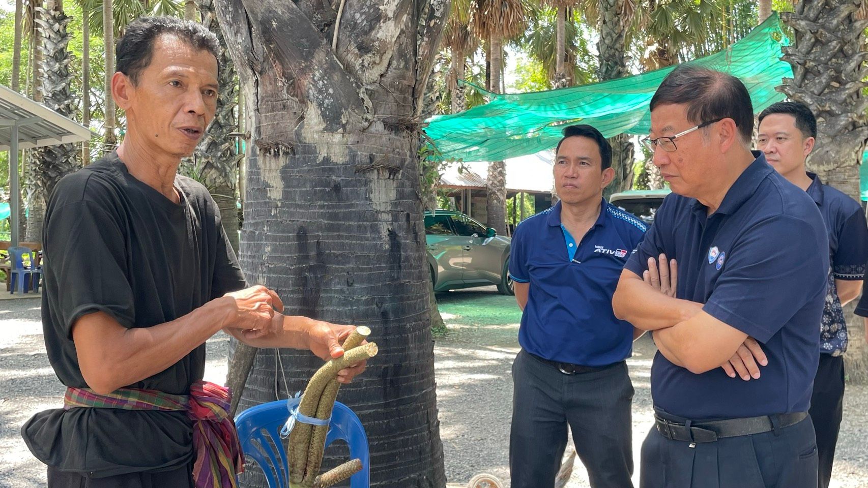 A local artisan demonstrates natural fiber craftwork to two officials outdoors under trees.