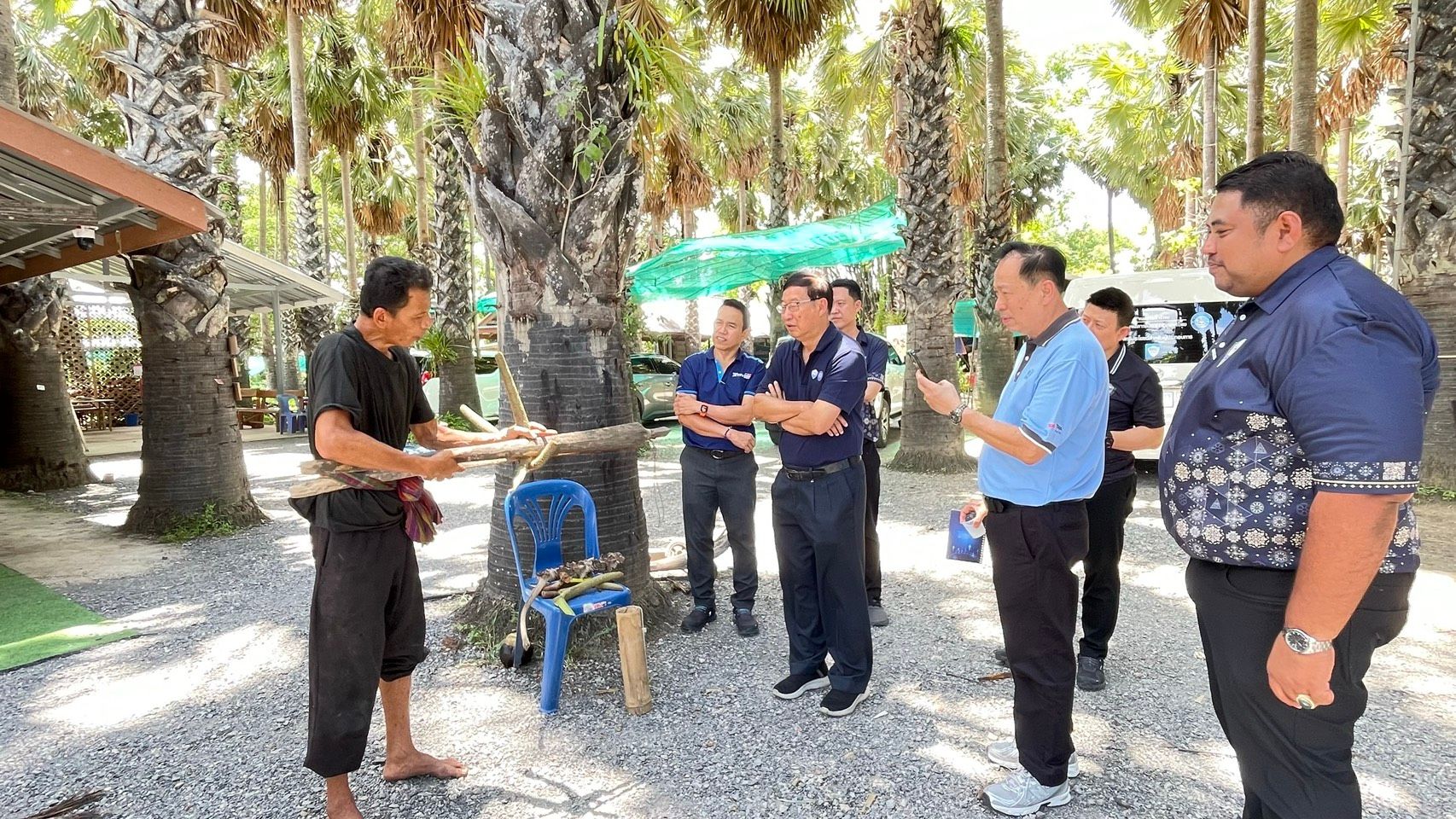 Men discuss in a palm grove, one holding traditional wooden tools.