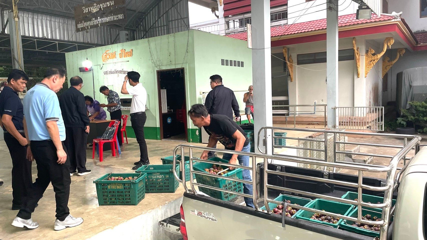 Men load green crates of produce into a pickup truck at an outdoor market.