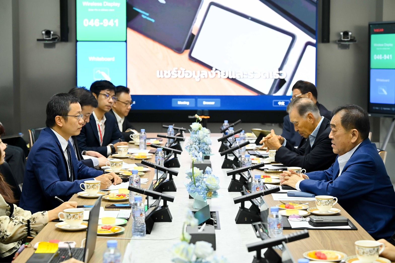 Men in suits at a conference table with a large screen displaying digital content.