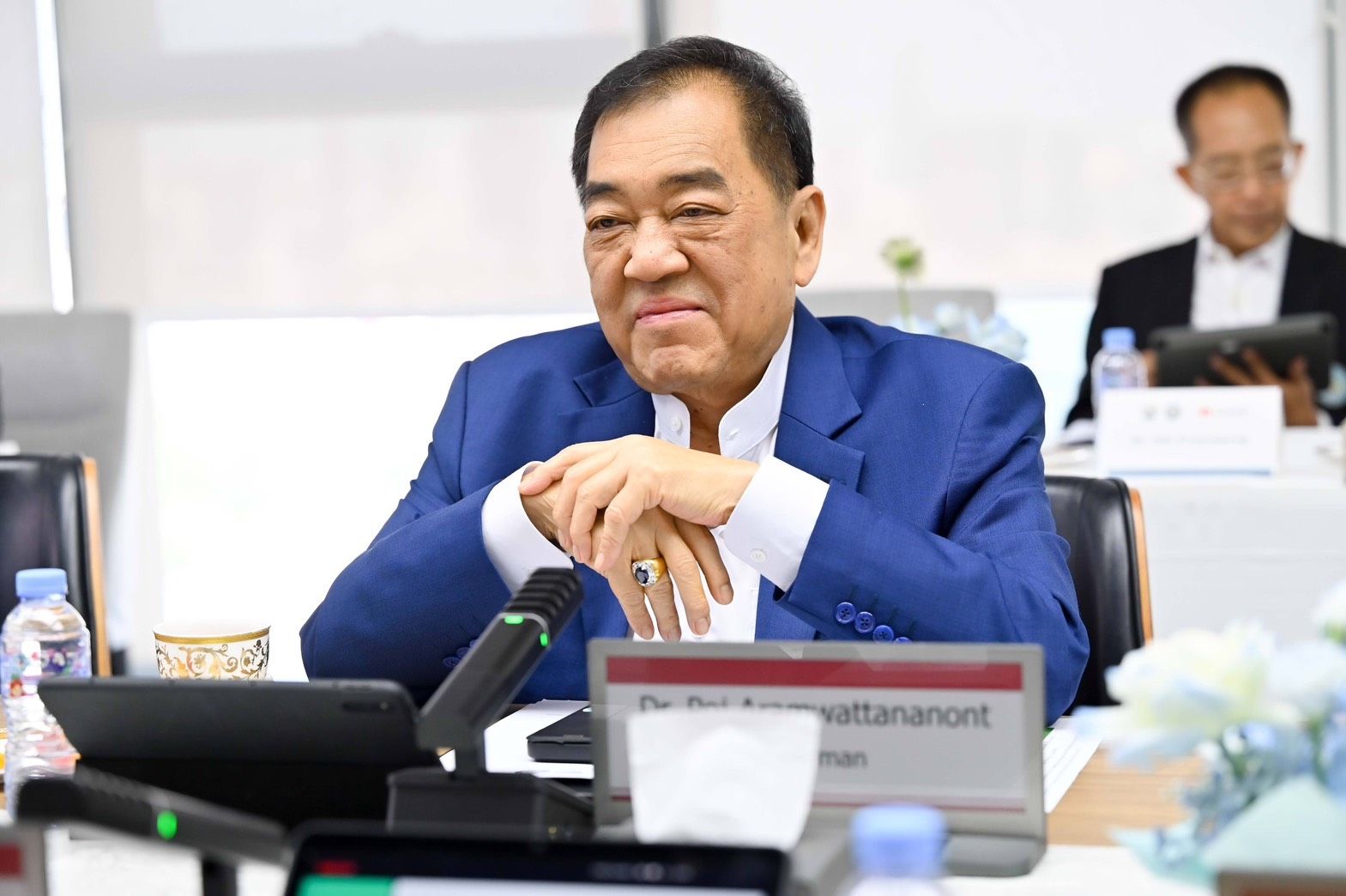 A distinguished man in a blue suit sits at a conference table with clasped hands.