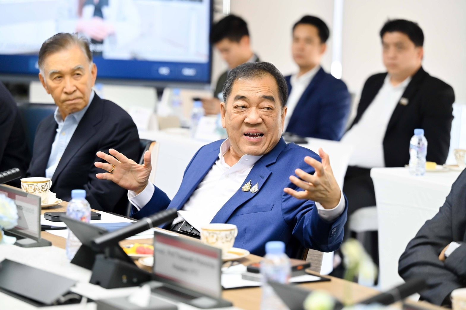 Man in blue jacket actively speaking, gesturing at a conference table with colleagues.