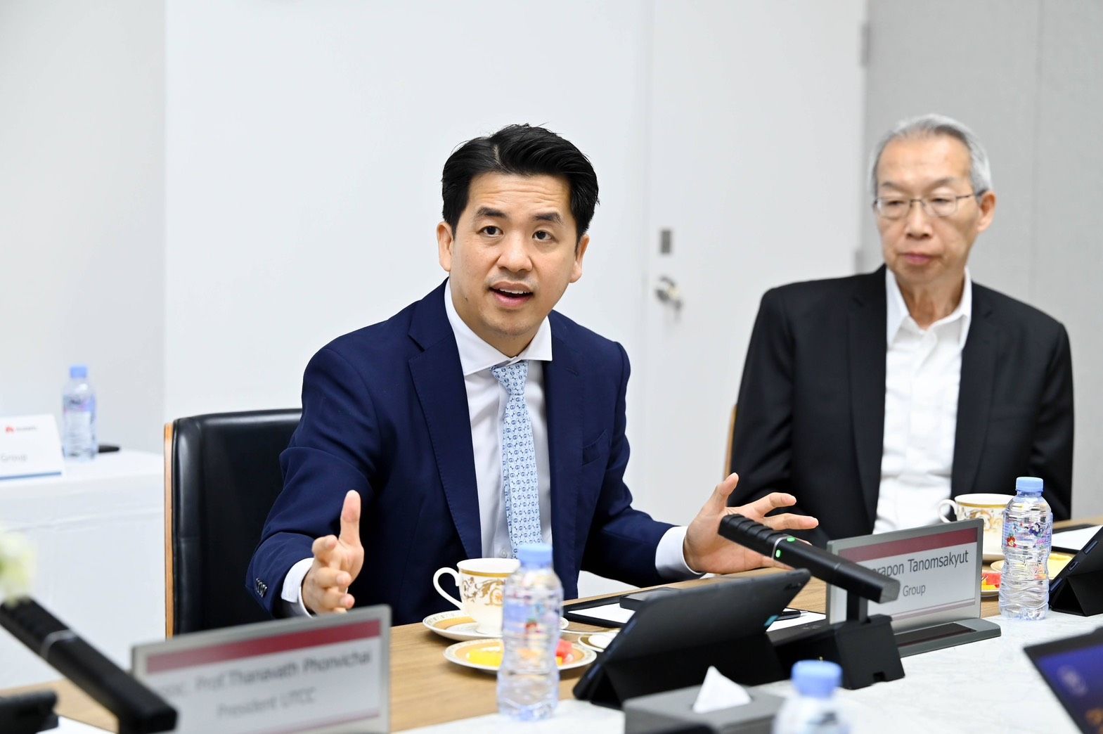 A man in a blue suit speaks animatedly at a conference table with another man.
