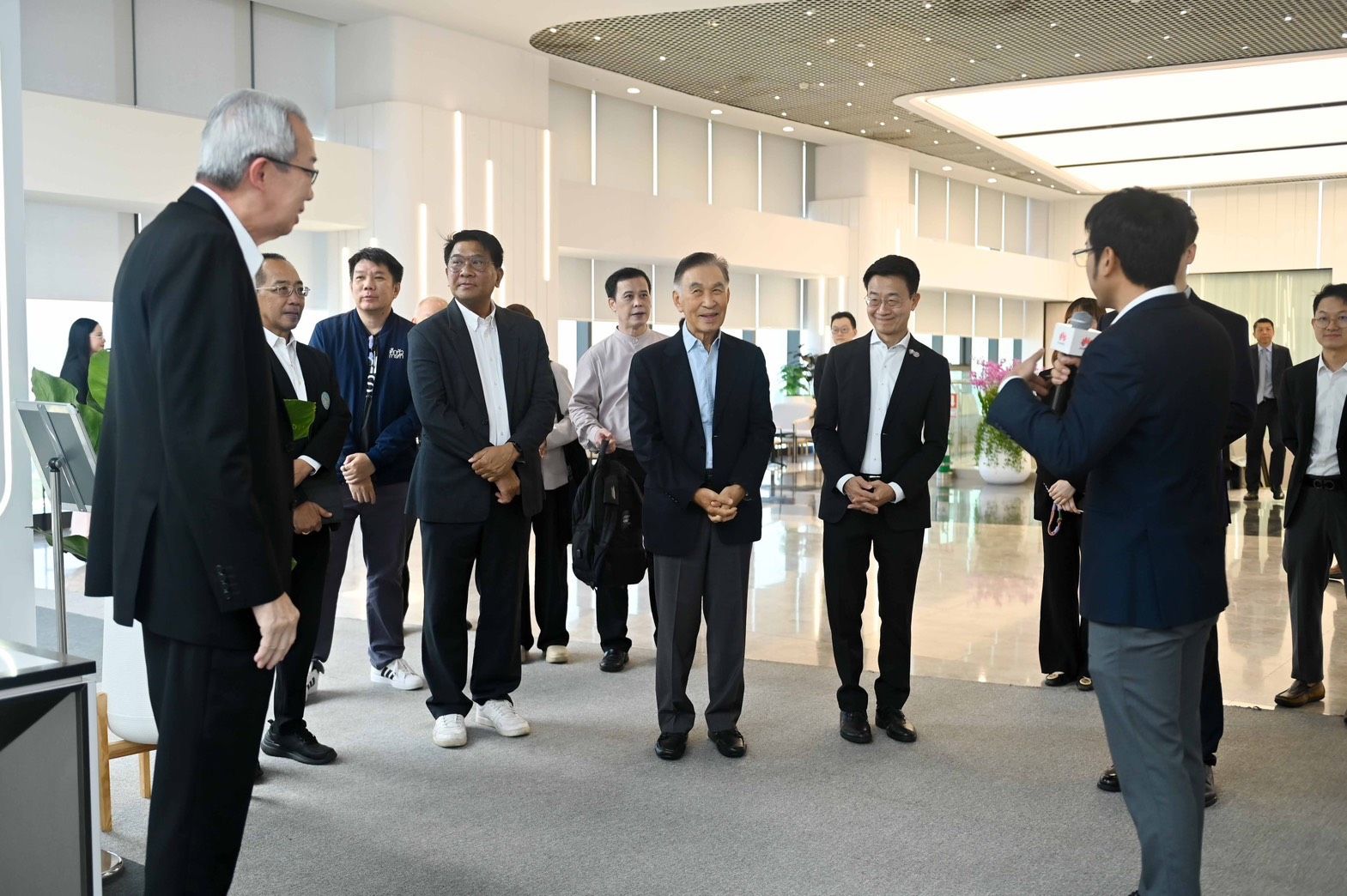 A group of businessmen in suits attentively listening to a speaker in a modern office lobby.