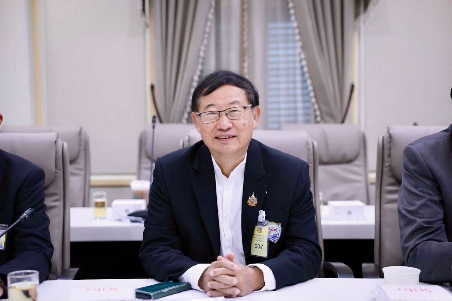 A man in a dark suit and glasses smiles while sitting at a conference table.
