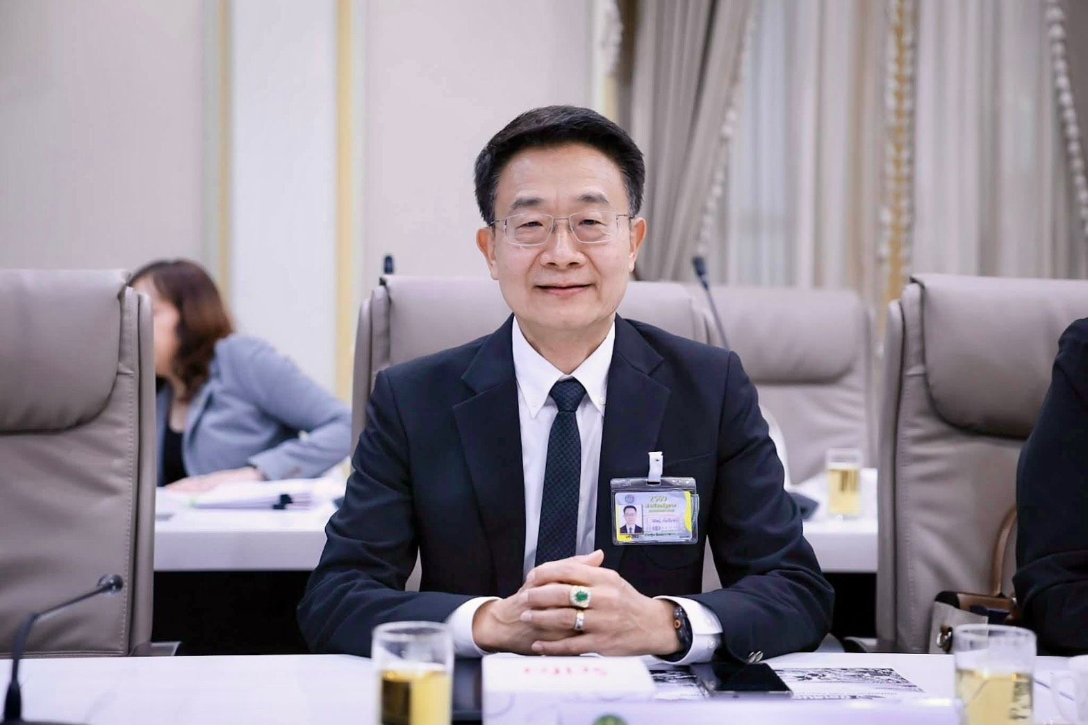 East Asian man in a dark suit and glasses smiles, seated at a meeting table.