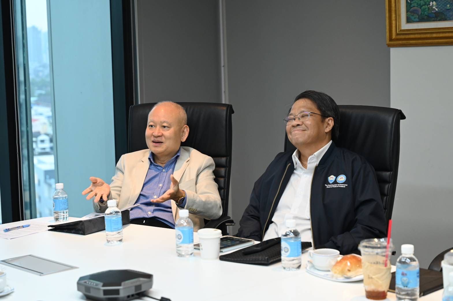 Two men sit at a modern conference table, one gesturing while the other smiles.