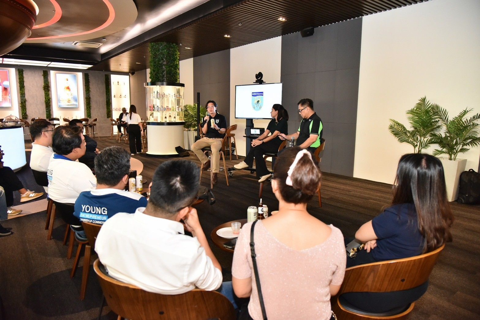 Attendees listen to three speakers presenting on a stage in a modern cafe setting.