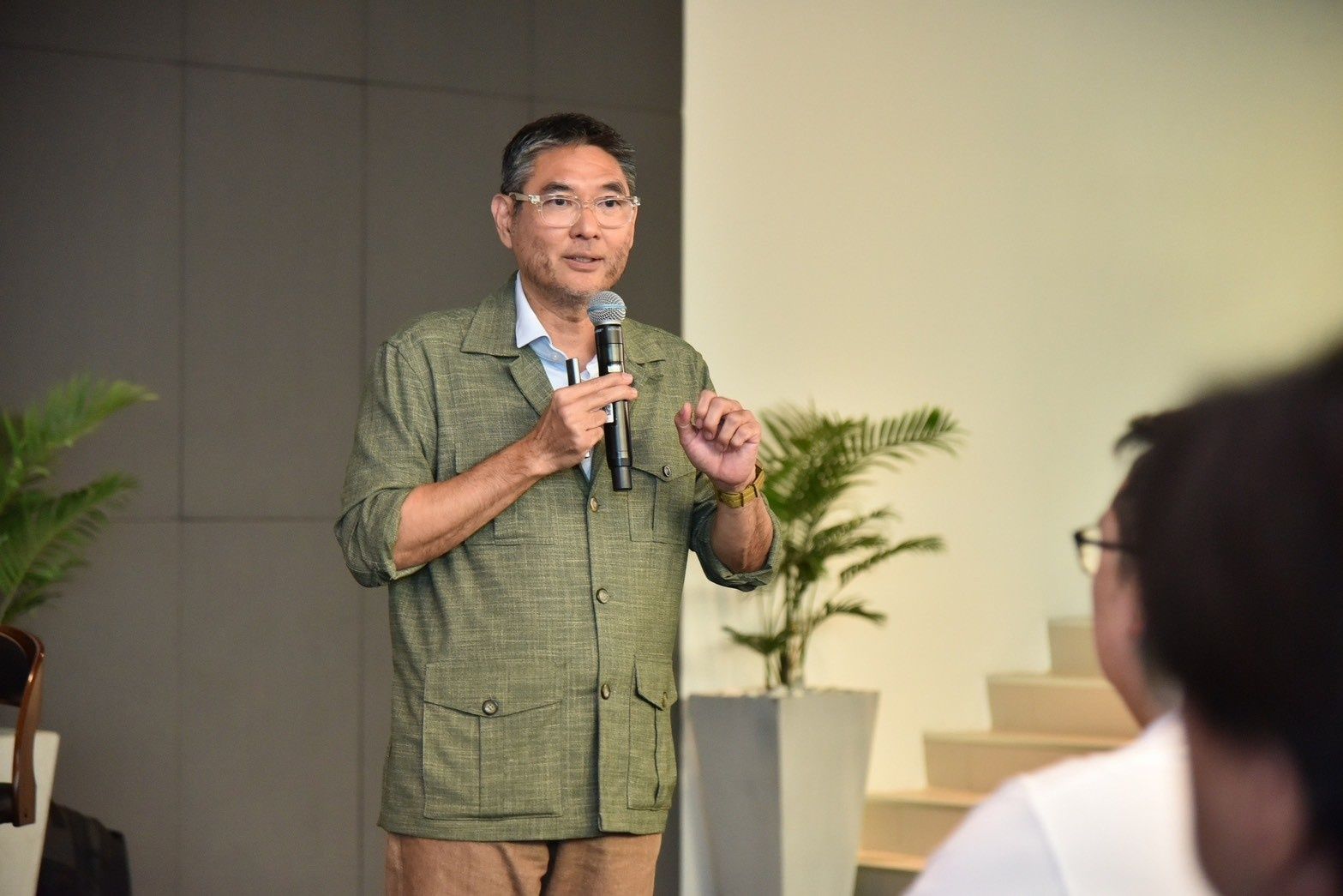 Man in green jacket speaks into microphone, gesturing during a presentation.