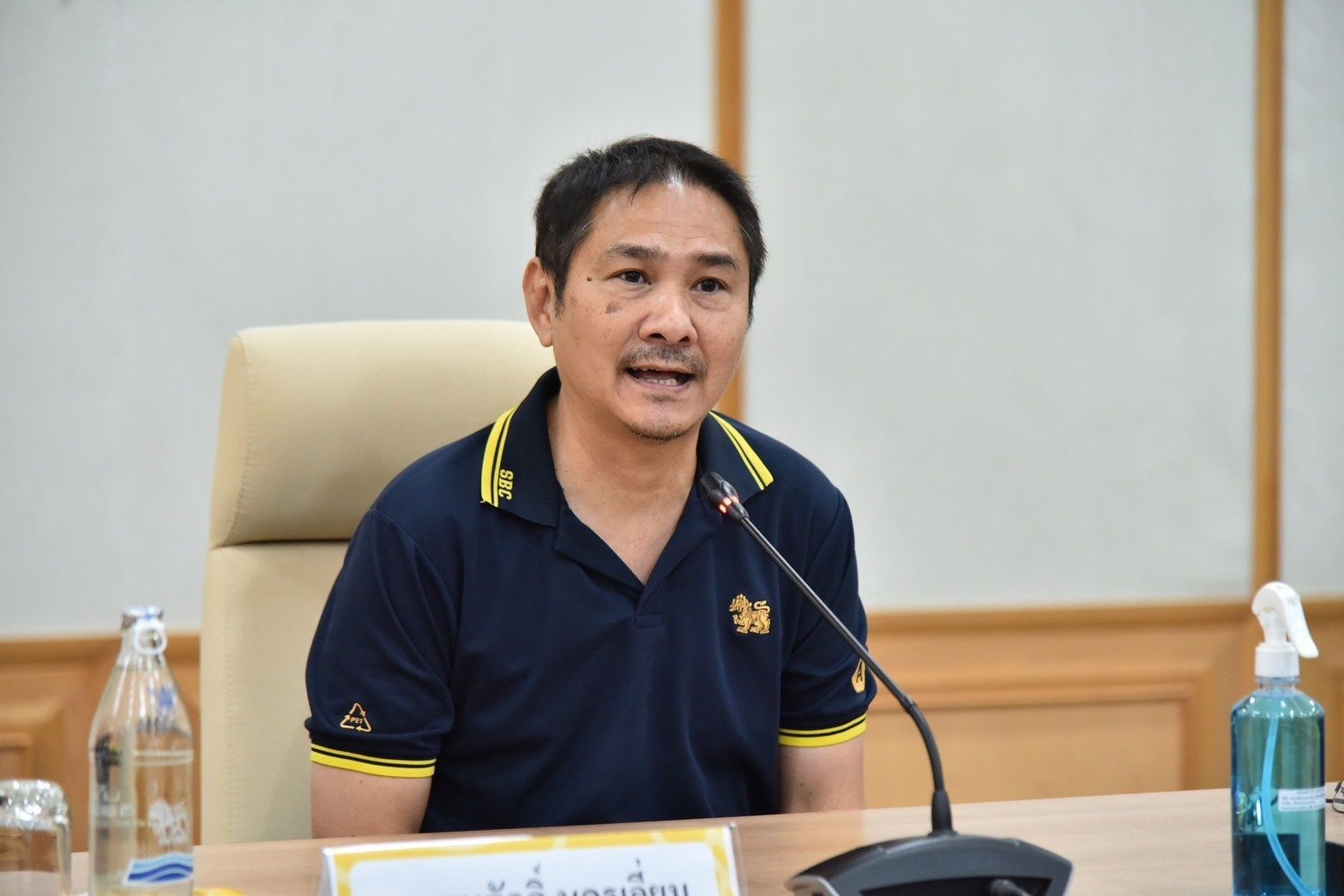 Man in blue polo shirt speaking into a microphone at a meeting table.