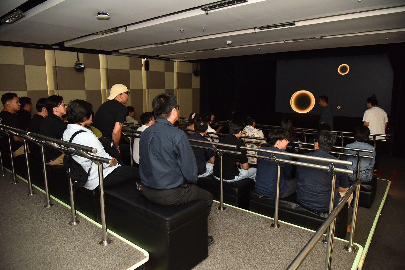 Audience seated in dark room watching glowing rings on a large screen.