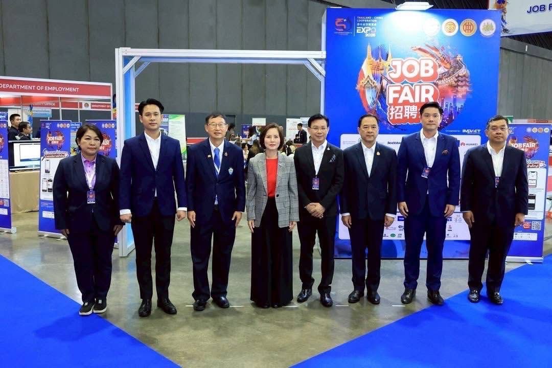 Seven professionals stand together in formal attire at a bustling job fair event.