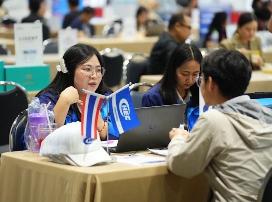 Woman in glasses speaks to a man at a table with flags and a laptop.