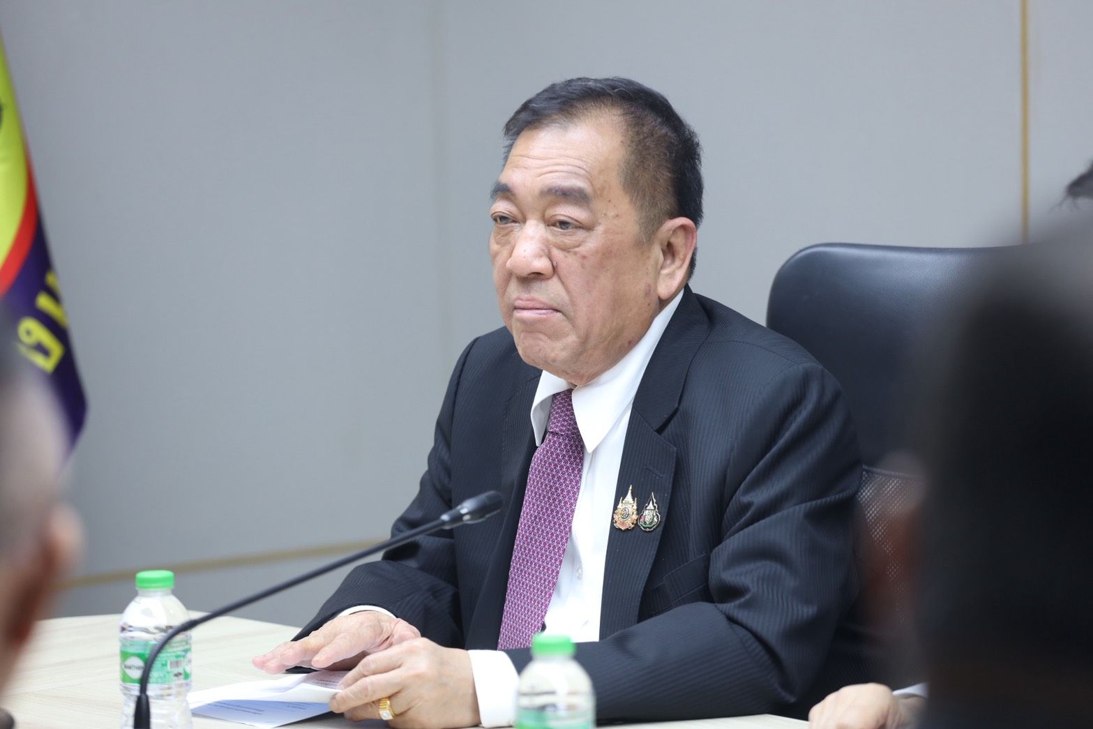 Older Asian man in a dark suit and purple tie sits seriously at a meeting table.