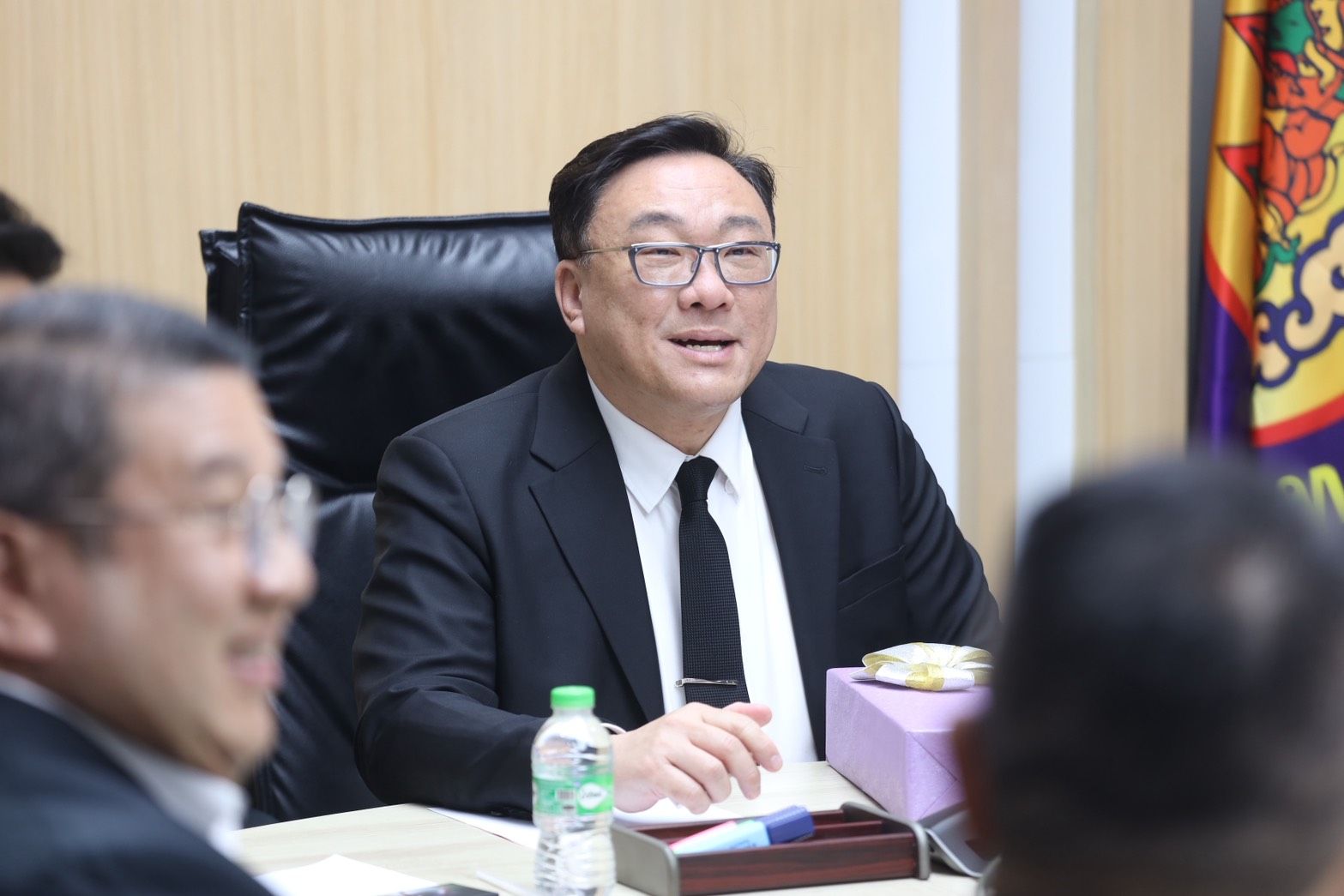 A man in a dark suit and glasses speaks animatedly at a conference table.