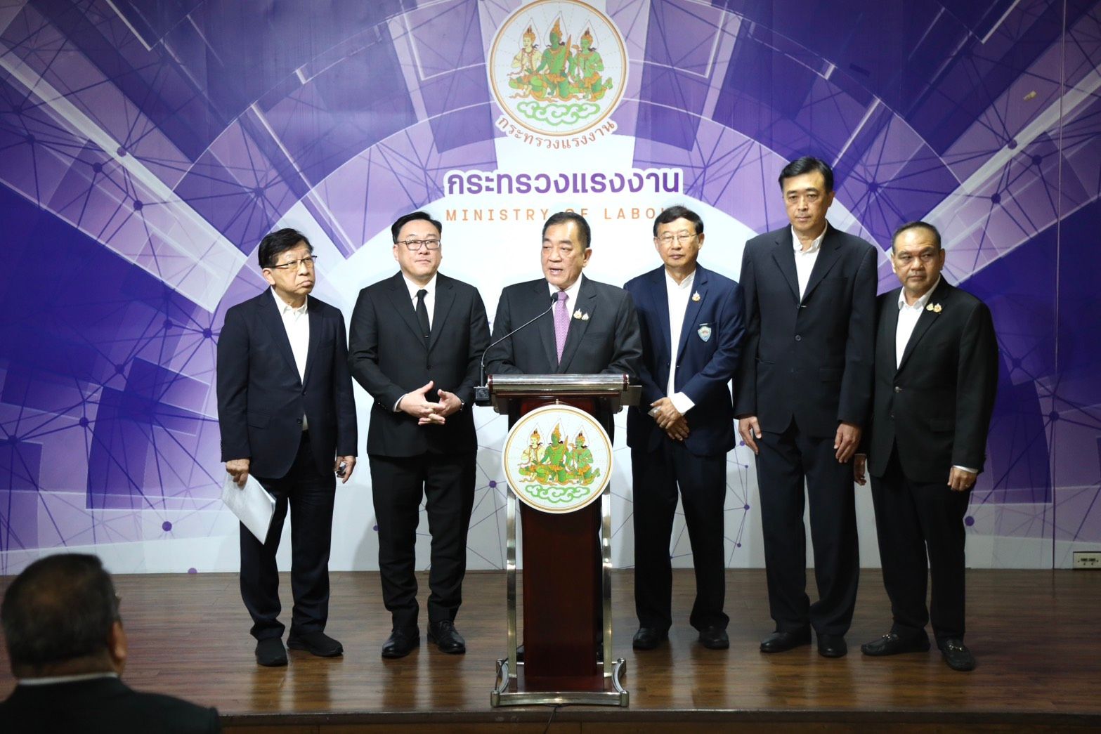 Six men in suits stand behind a podium with the Ministry of Labour logo.