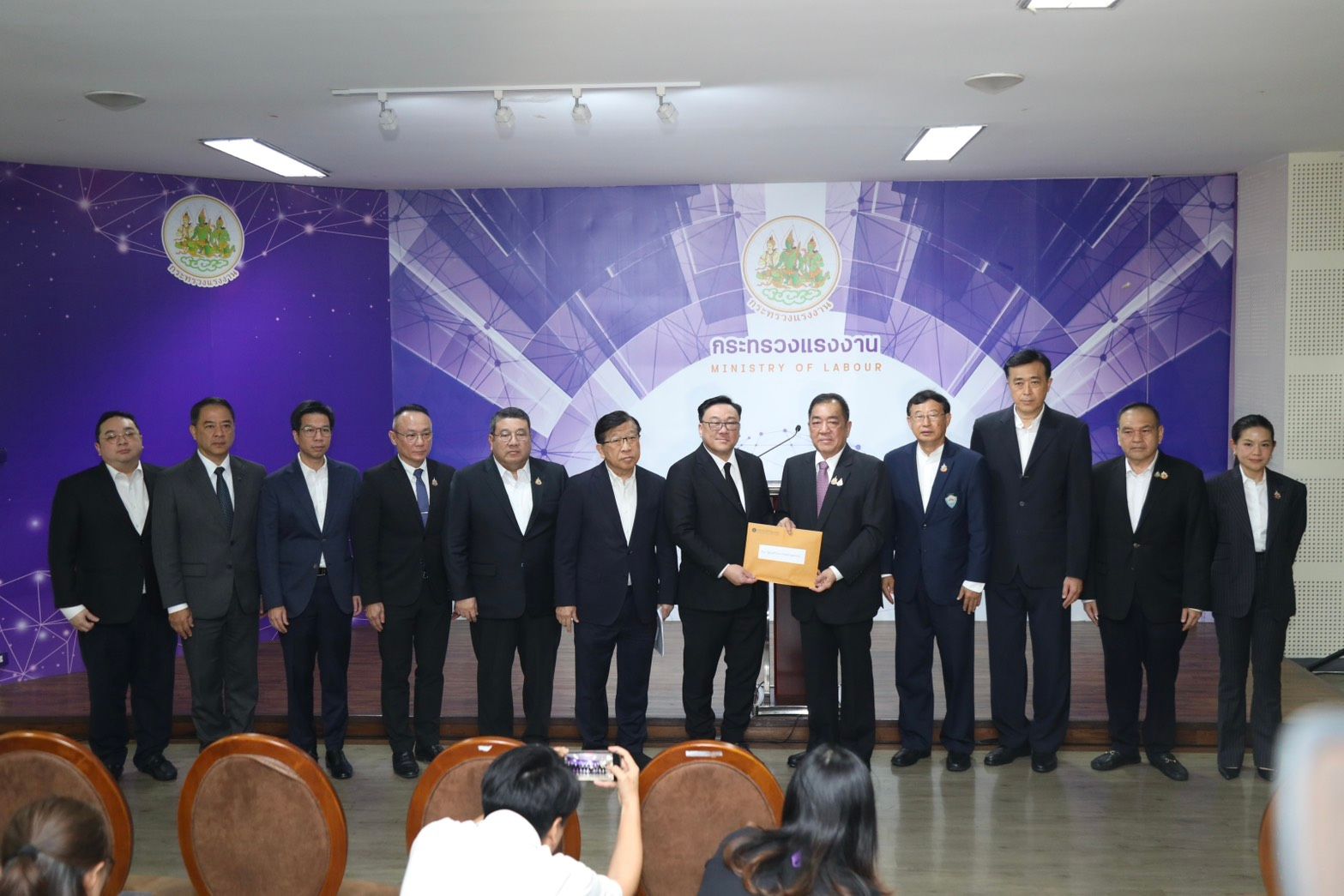 Formally dressed men stand before a purple banner as one receives a golden plaque.