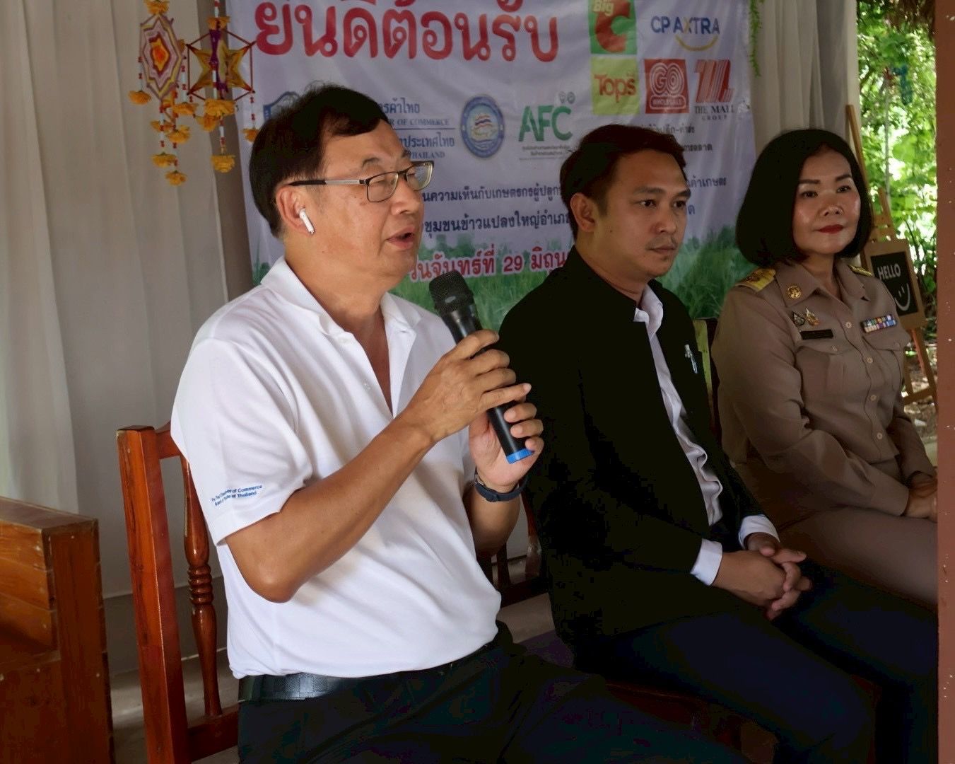 Man in glasses speaks into microphone, seated with two officials at an event.