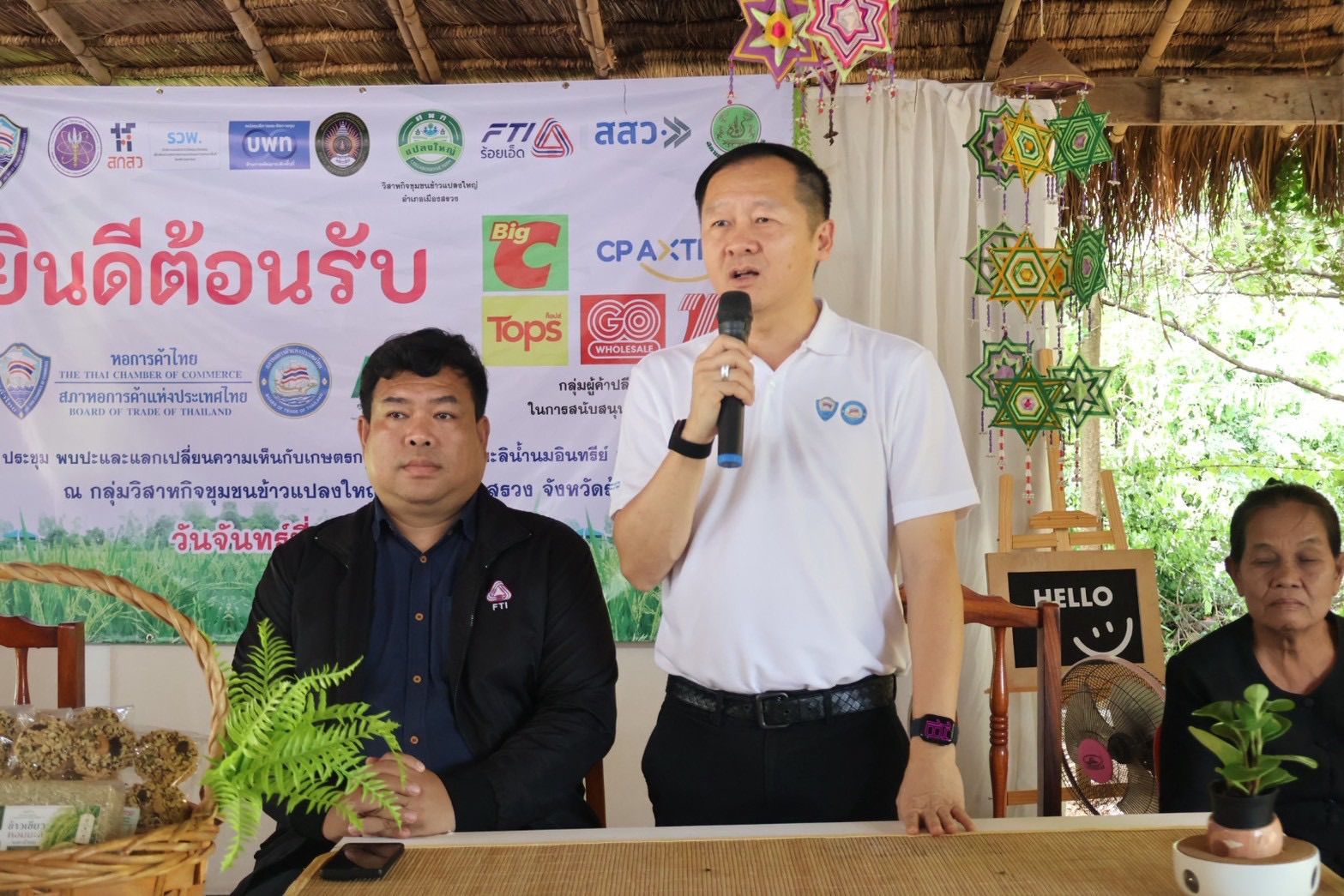 Man in white polo shirt speaks into microphone at outdoor event with welcome banner.