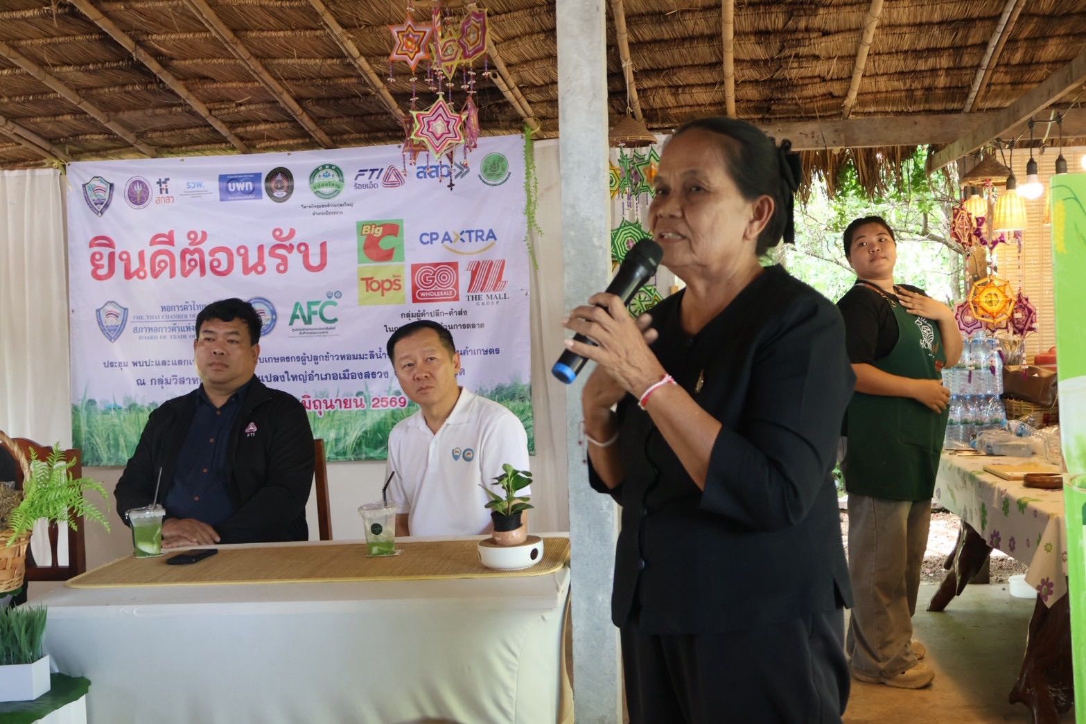 A woman speaks into a microphone while three men sit at a table in the background.