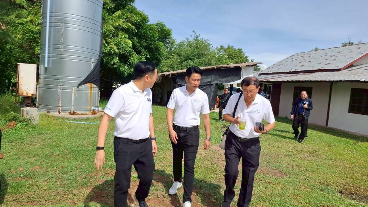 Three men walk past a large silver silo and building on a grassy path outdoors.