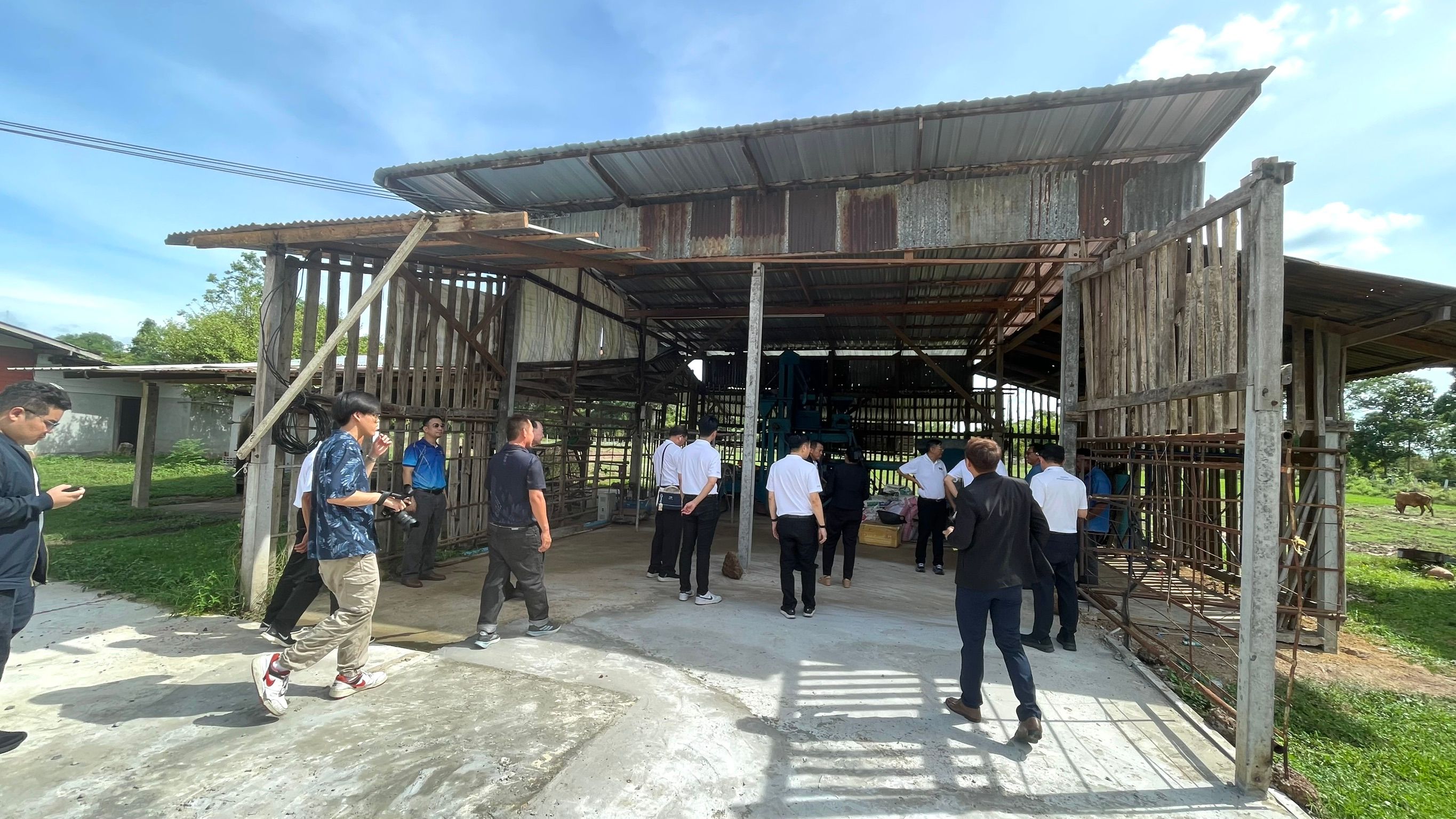Group of people gathered near and inside an old, open-sided barn with a metal roof.